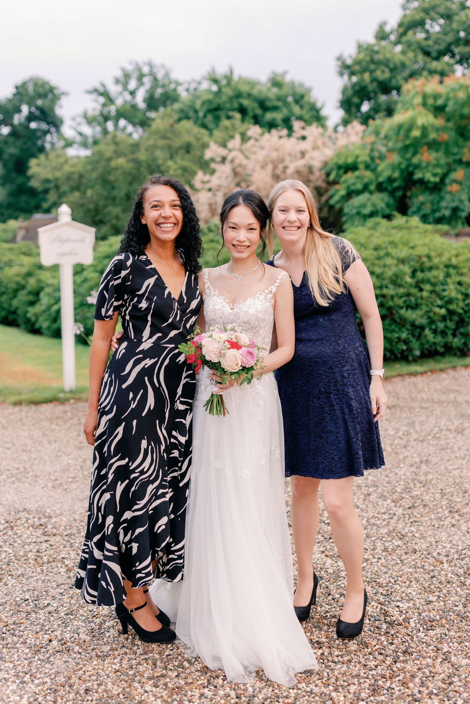Three women pose together on a gravel path, with one wearing a white wedding gown holding a pink bouquet.