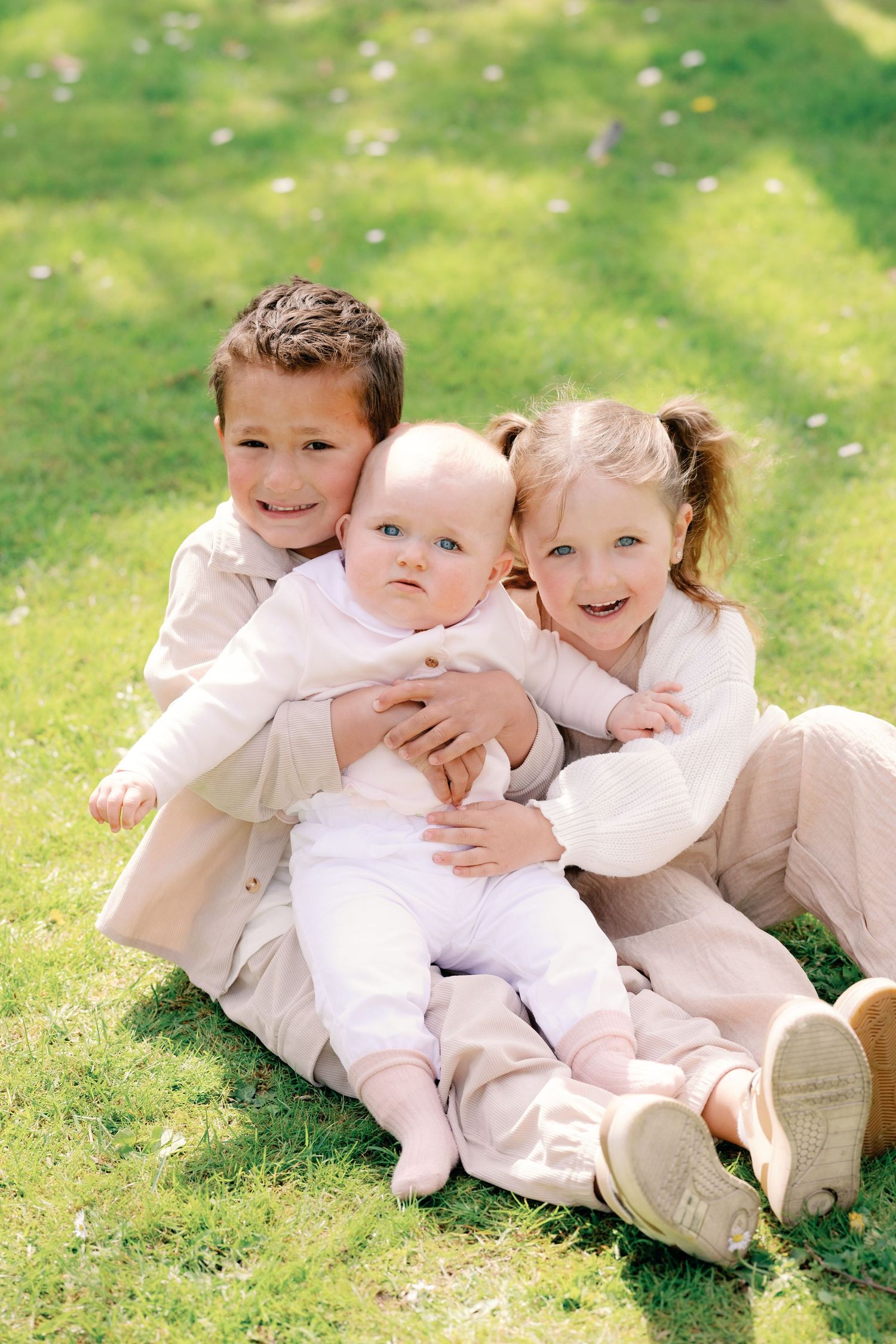 Two children and a baby sitting on green grass outdoors, smiling happily together on a sunny day.