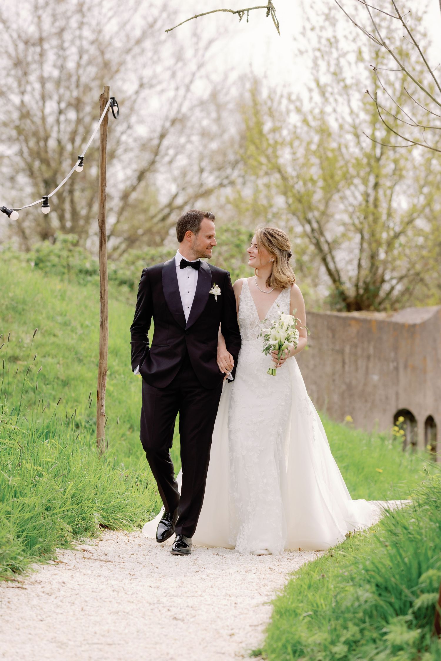 Bride and groom walk hand in hand on a garden path, she holds a bouquet, he wears a black tuxedo.