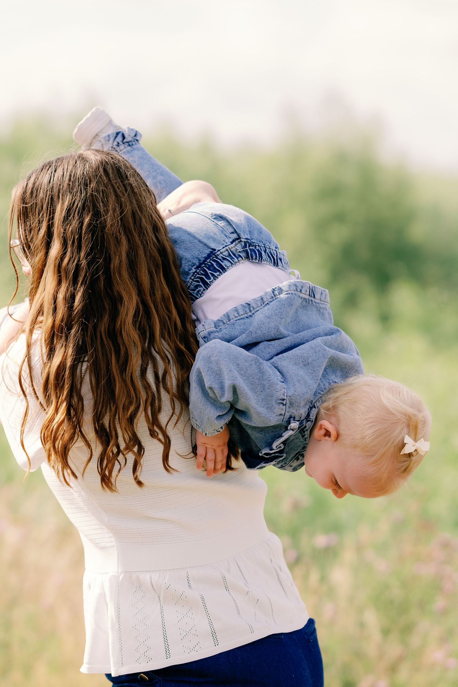 A mother playfully holds her toddler upside down outdoors, both wearing denim in a lush green field.
