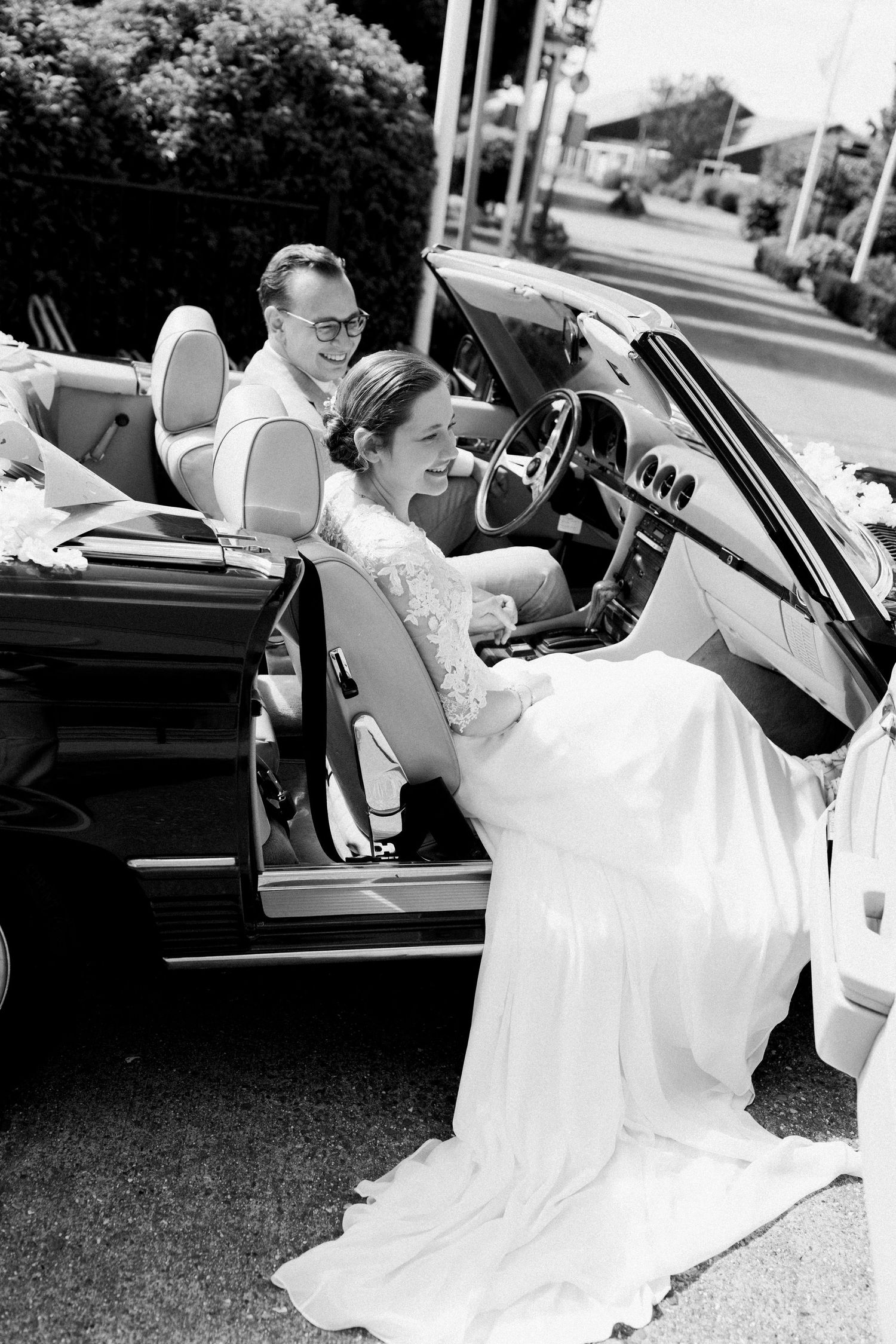 Black and white photo of a smiling bride in white gown getting into a classic convertible car with groom seated inside.
