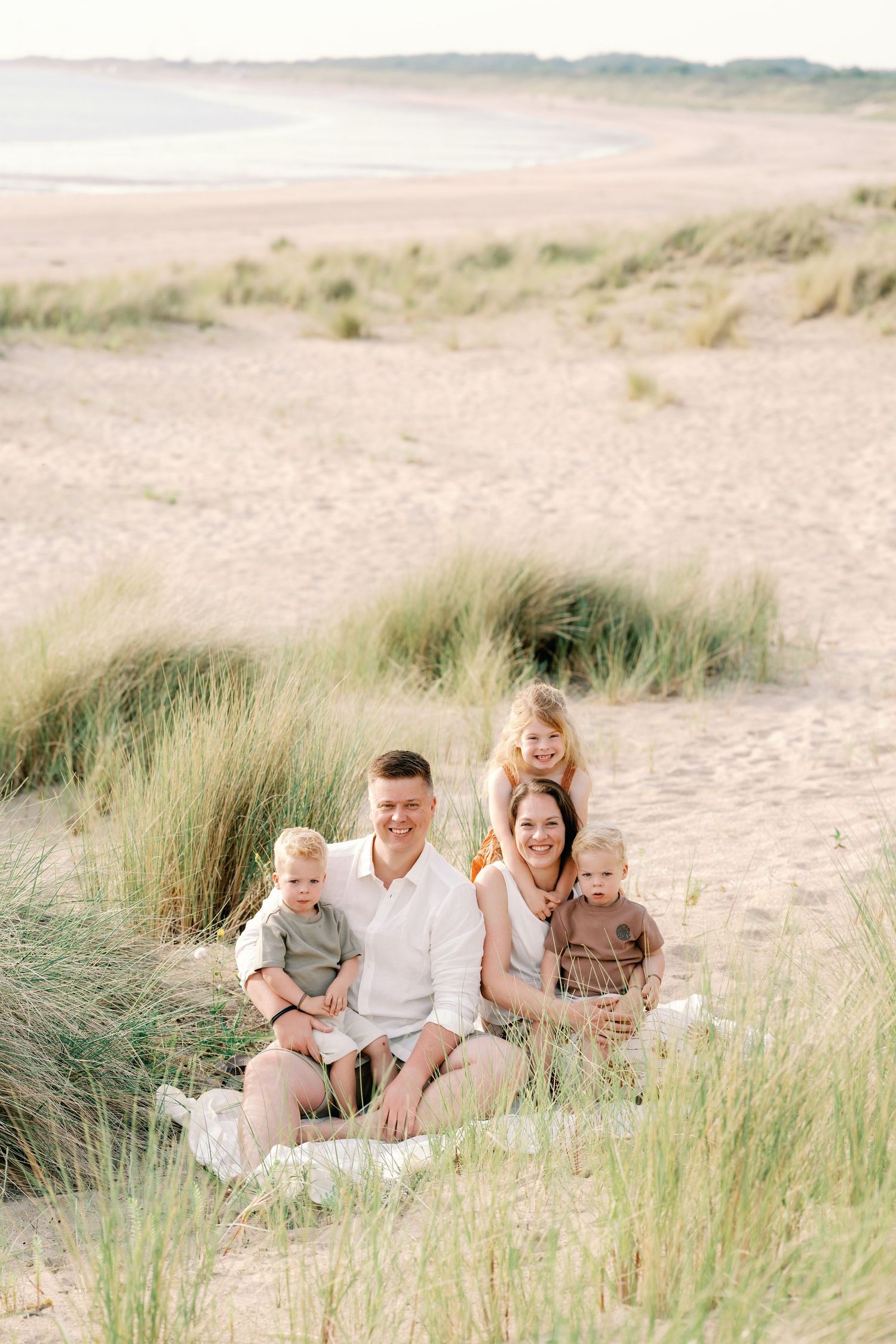 Family of five sitting on sandy beach dunes surrounded by grass, smiling at camera on a sunny day.