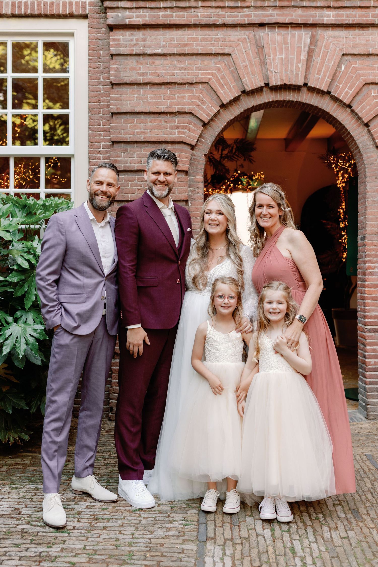 Wedding party poses in front of brick archway entrance with flower girls in white dresses and adults in formal attire.