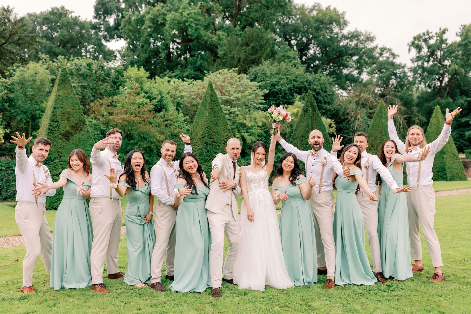 Wedding party in soft mint green and neutral attire poses joyfully on grass.
