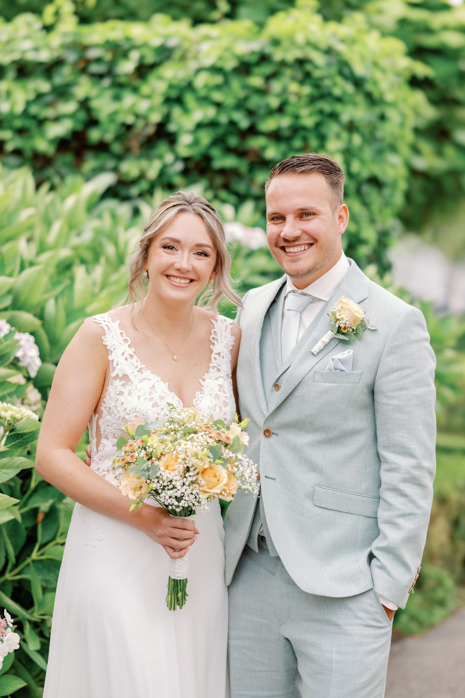 A romantic wedding portrait in a garden setting with the couple holding a peachy bouquet and wearing light gray attire.