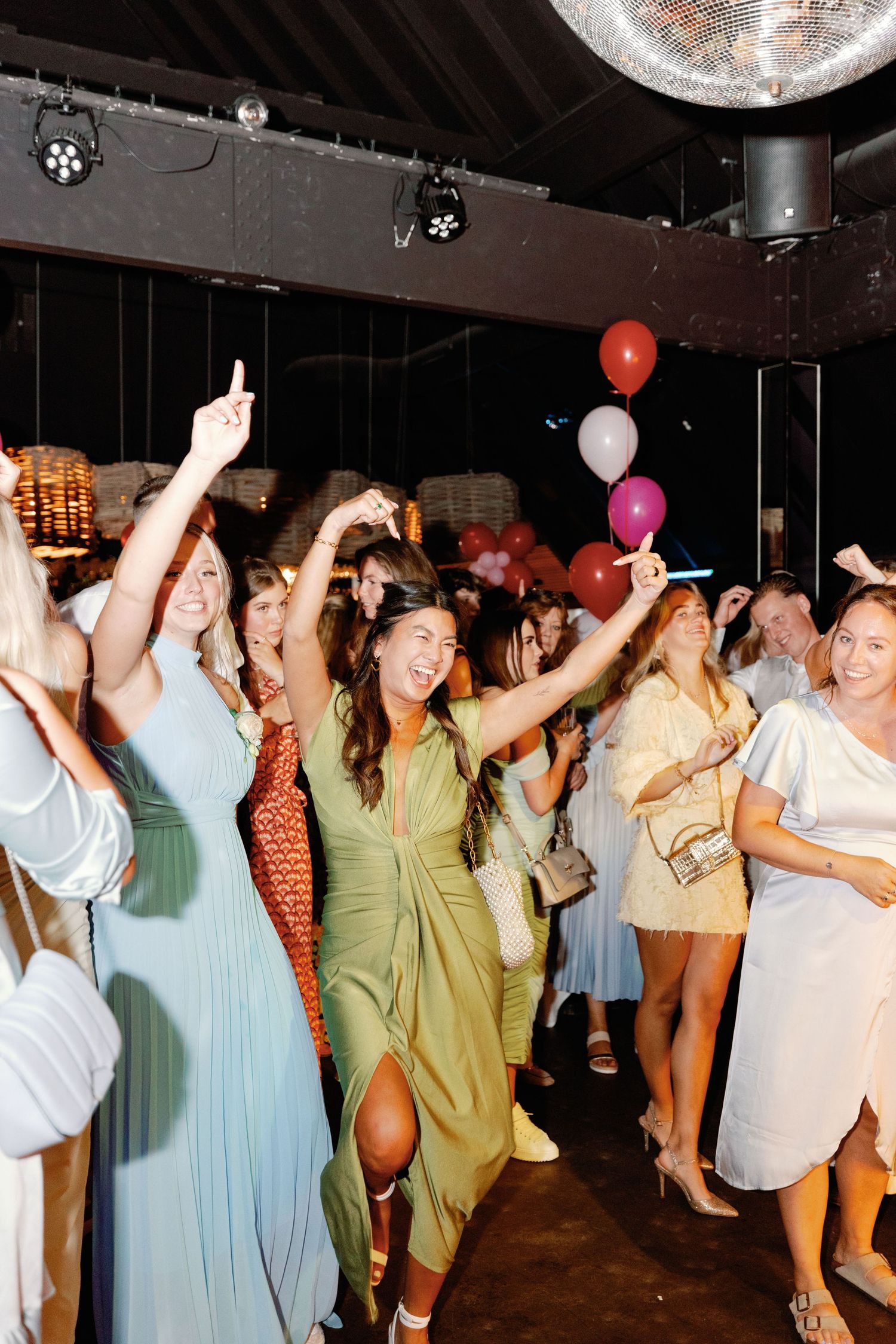 A group of party guests dancing and celebrating under disco balls with colorful balloons in a dark venue.