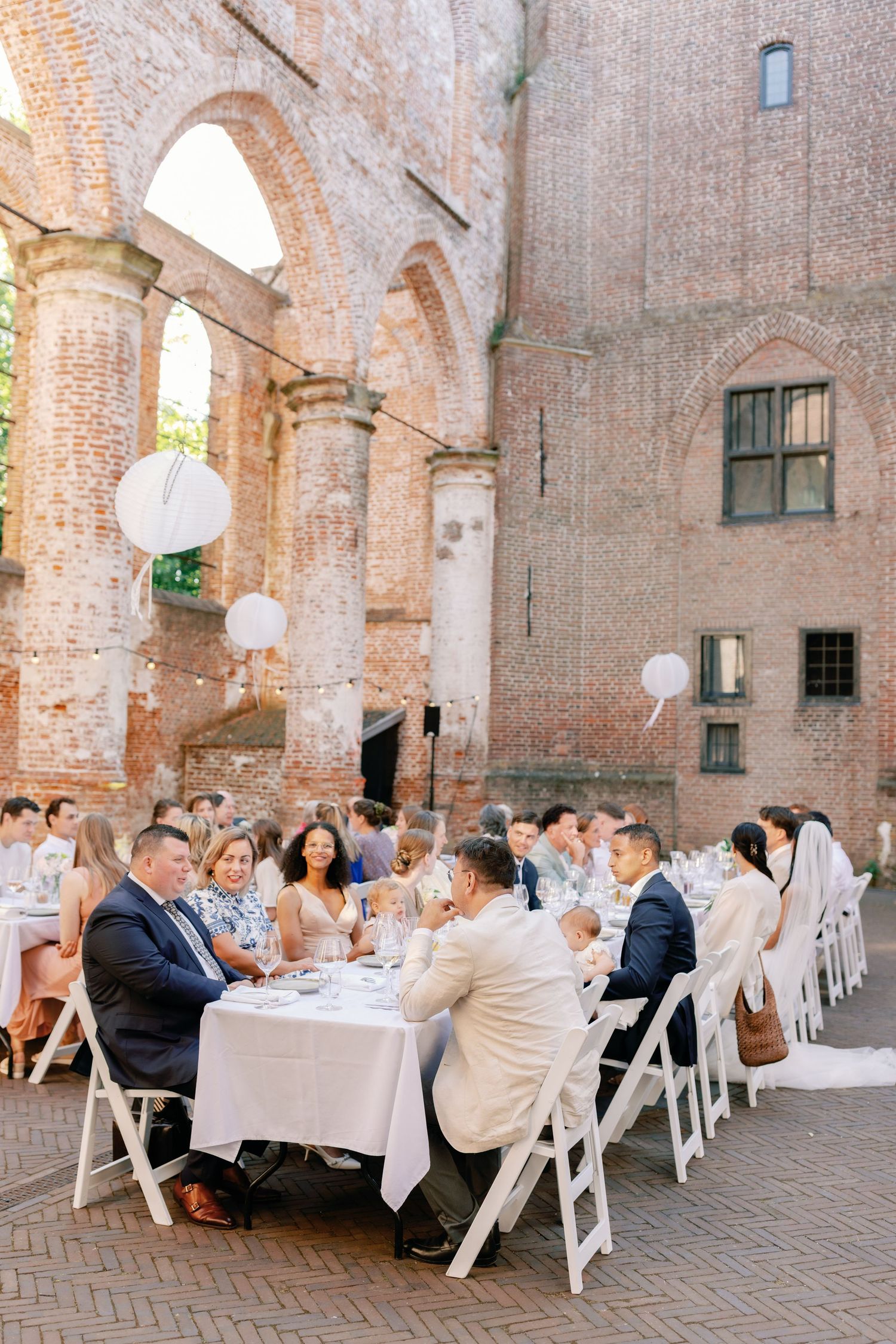 Elegant outdoor wedding reception dinner set in rustic brick ruins with white globe lanterns and folding chairs.