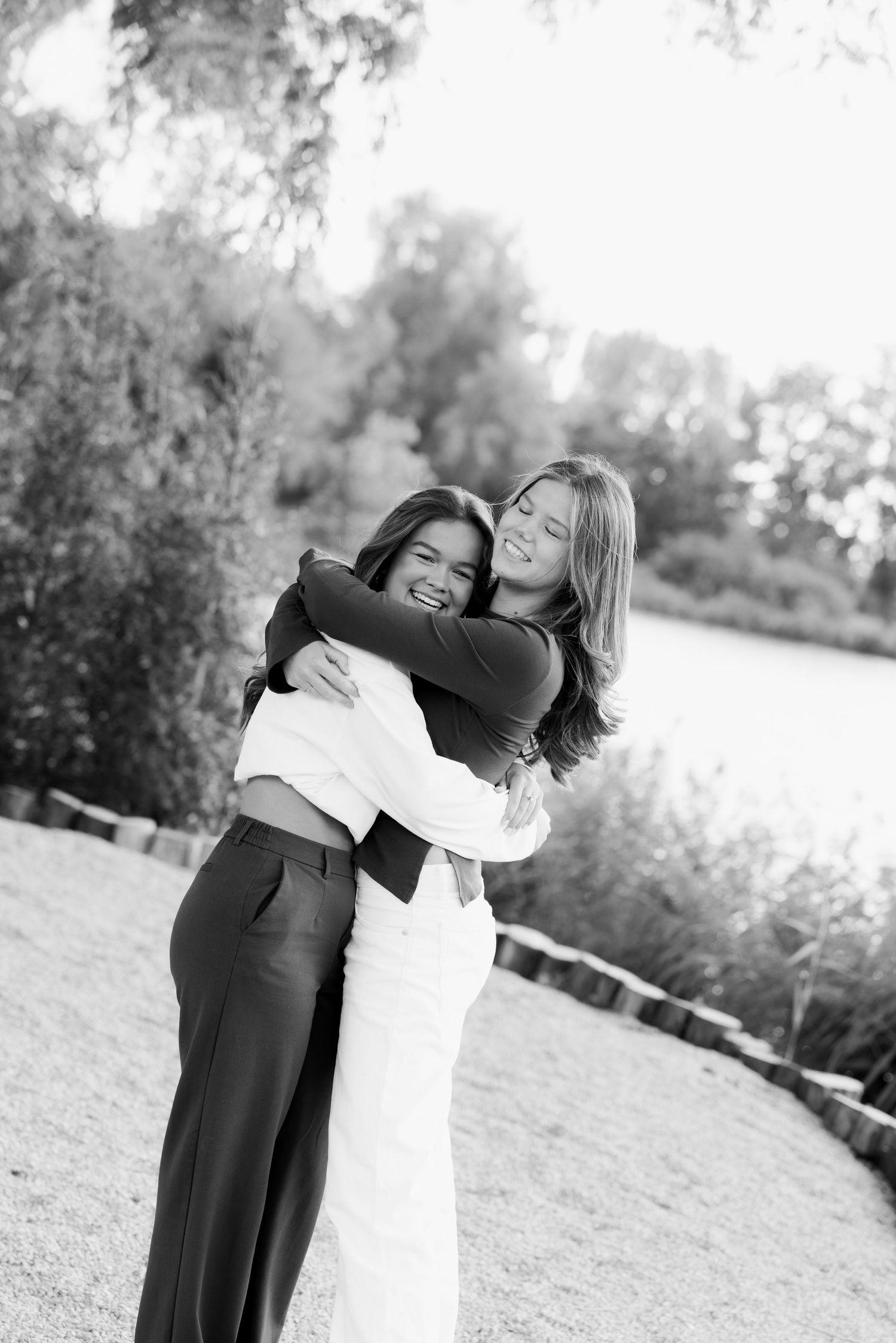 Two smiling young women embrace in a joyful hug outdoors in this black and white portrait, with a calm lake and trees in the background.