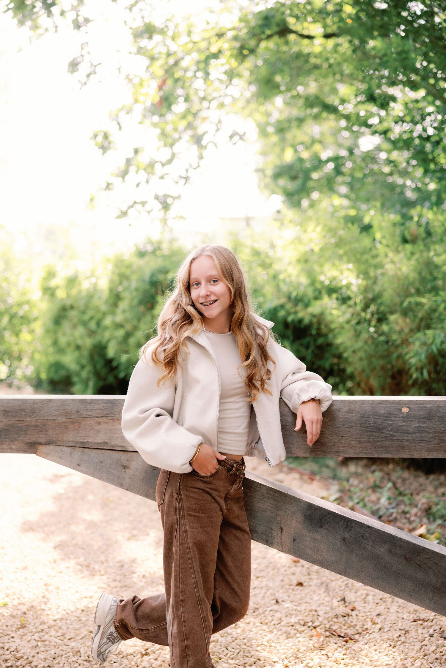 Smiling teenage girl with long wavy hair leans against a wooden fence outdoors in a cream jacket and brown corduroy pants among green trees.