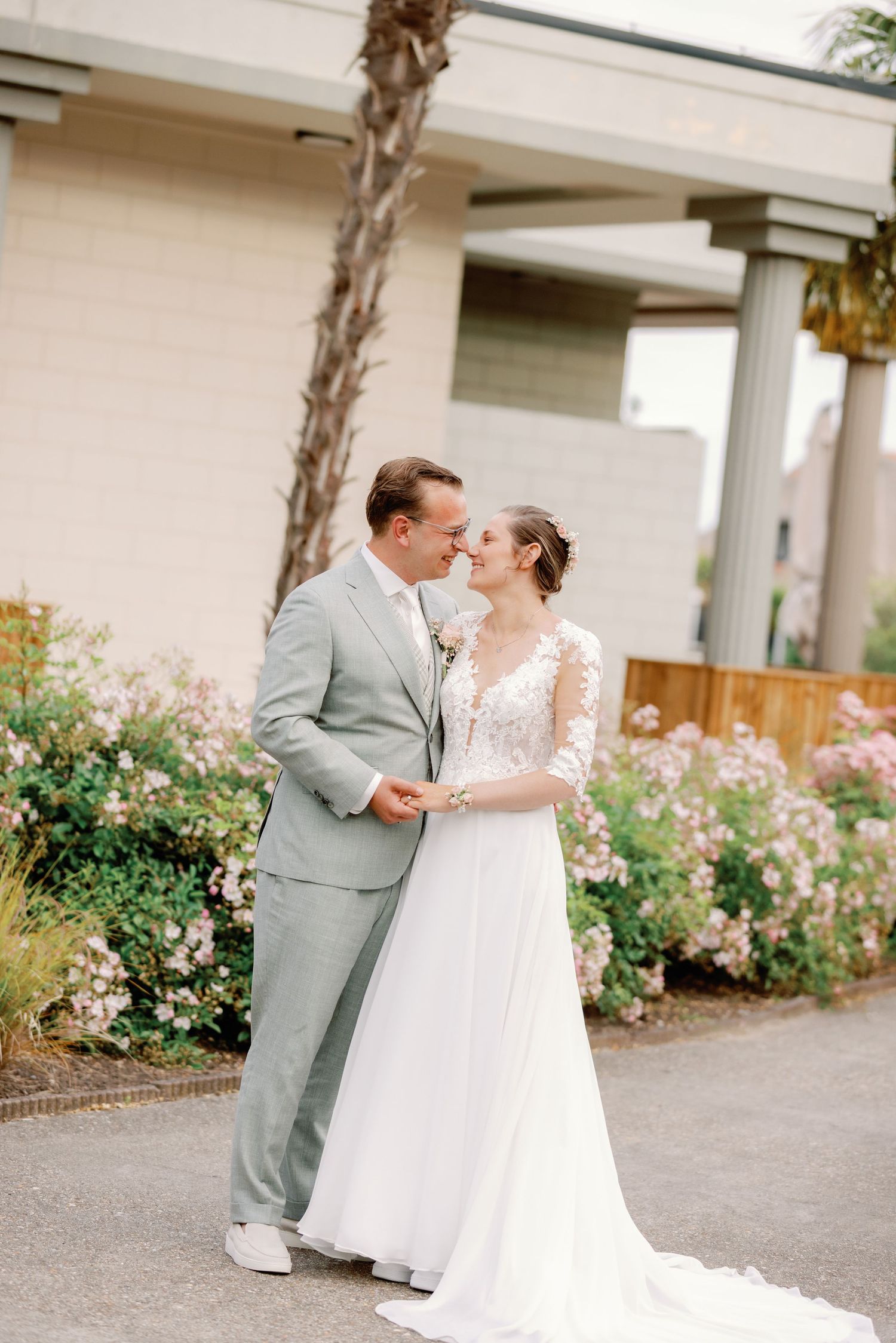 A bride and groom share a romantic moment outdoors, surrounded by blooming pink flowers, as she wears a lace gown and he a sage suit.