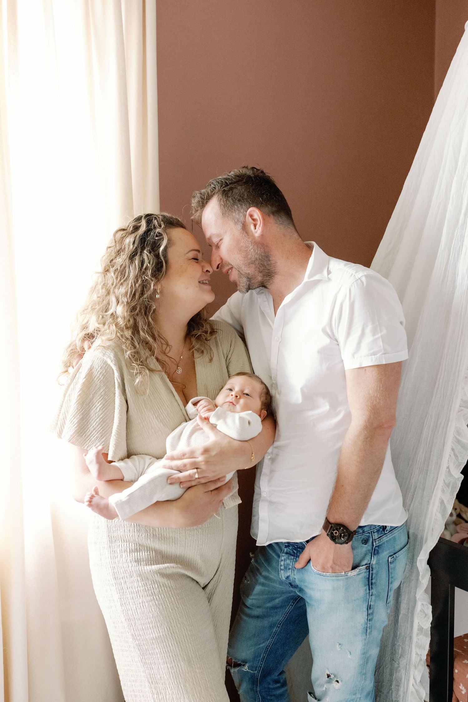 A couple in light colored clothing share intimate moments while holding their newborn.