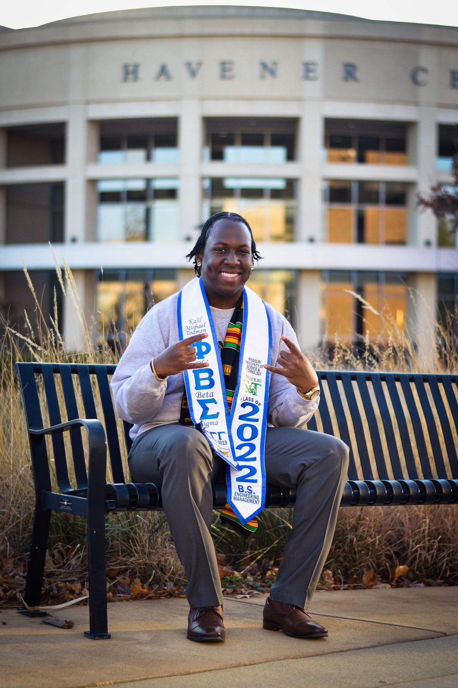 Graduate poses on bench outside Havener Center wearing academic regalia and blue graduation stole.