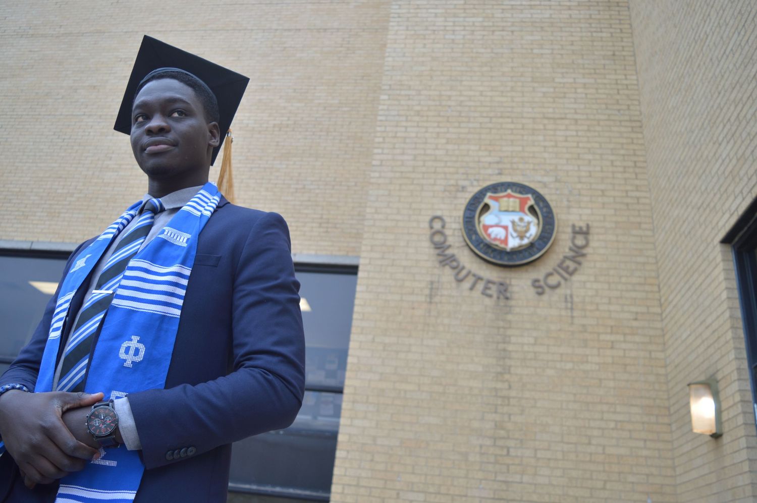 Black Graduate posing near university building entrance with stone facade.