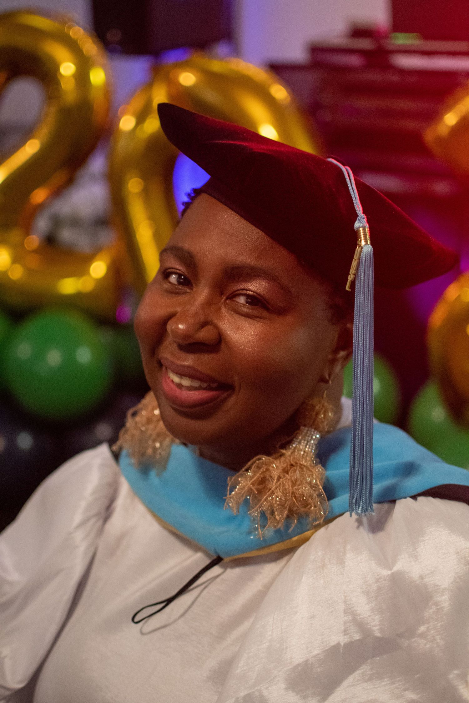Graduate wearing white cap and gown smiles during graduation ceremony with gold balloons in background.