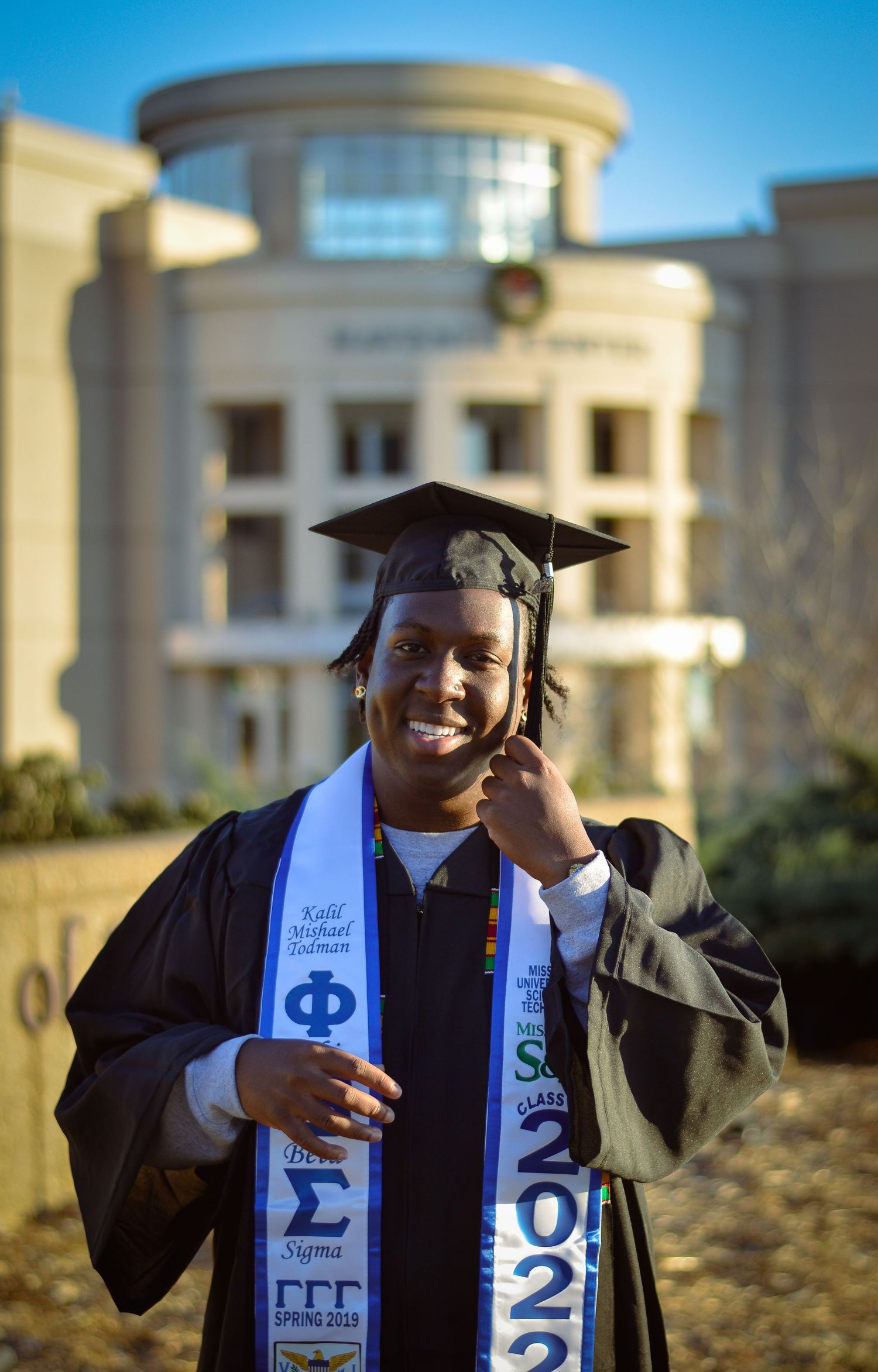 Graduate in cap and gown celebrating with diploma against architectural backdrop.
