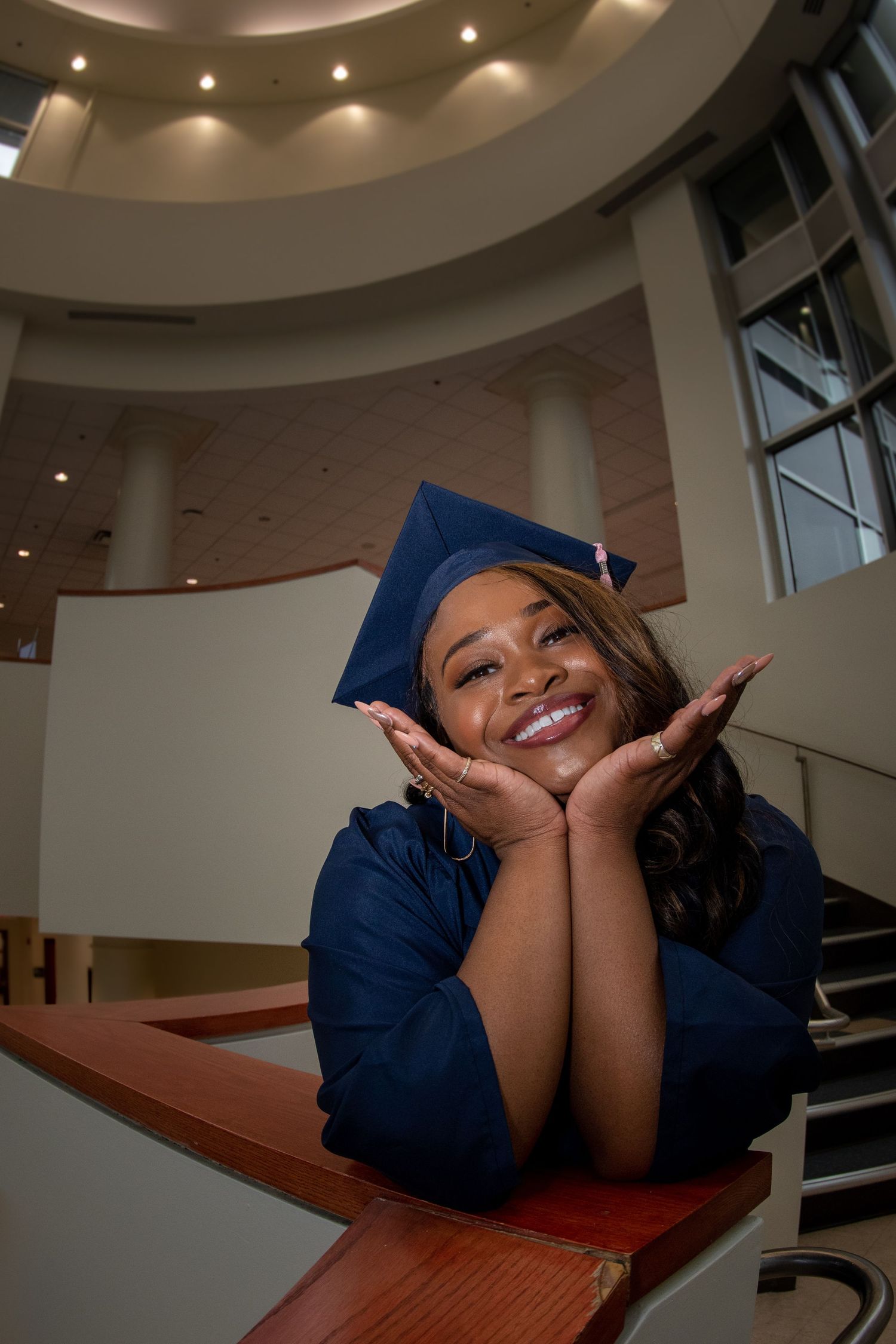 Graduate poses playfully on wooden desk during indoor photo session.