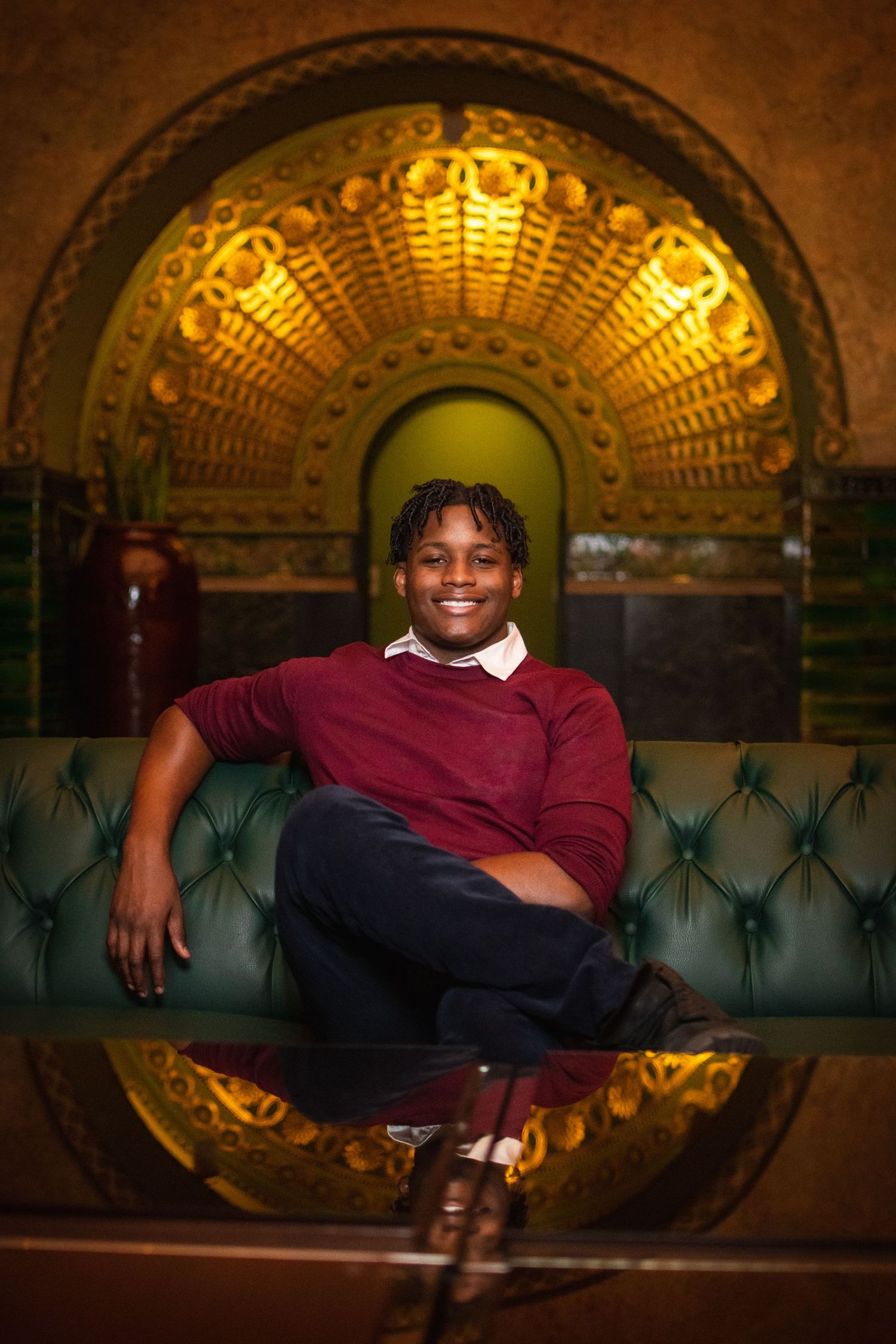 Artistic portrait in warm lighting against ornate gold architectural details and tufted green seating.