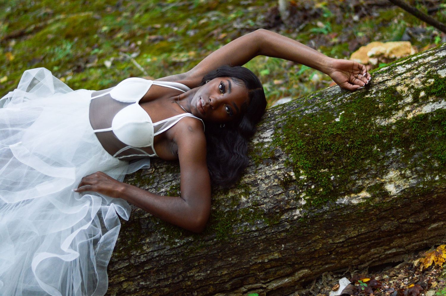 A figure in a flowing white dress poses gracefully on a fallen tree trunk in a lush forest setting.