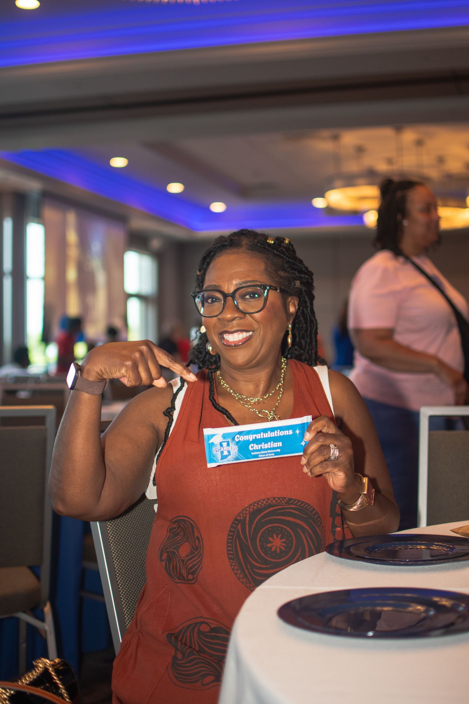 A person in a rust-colored tank top with circular designs smiles while seated at a table with blue lighting overhead.