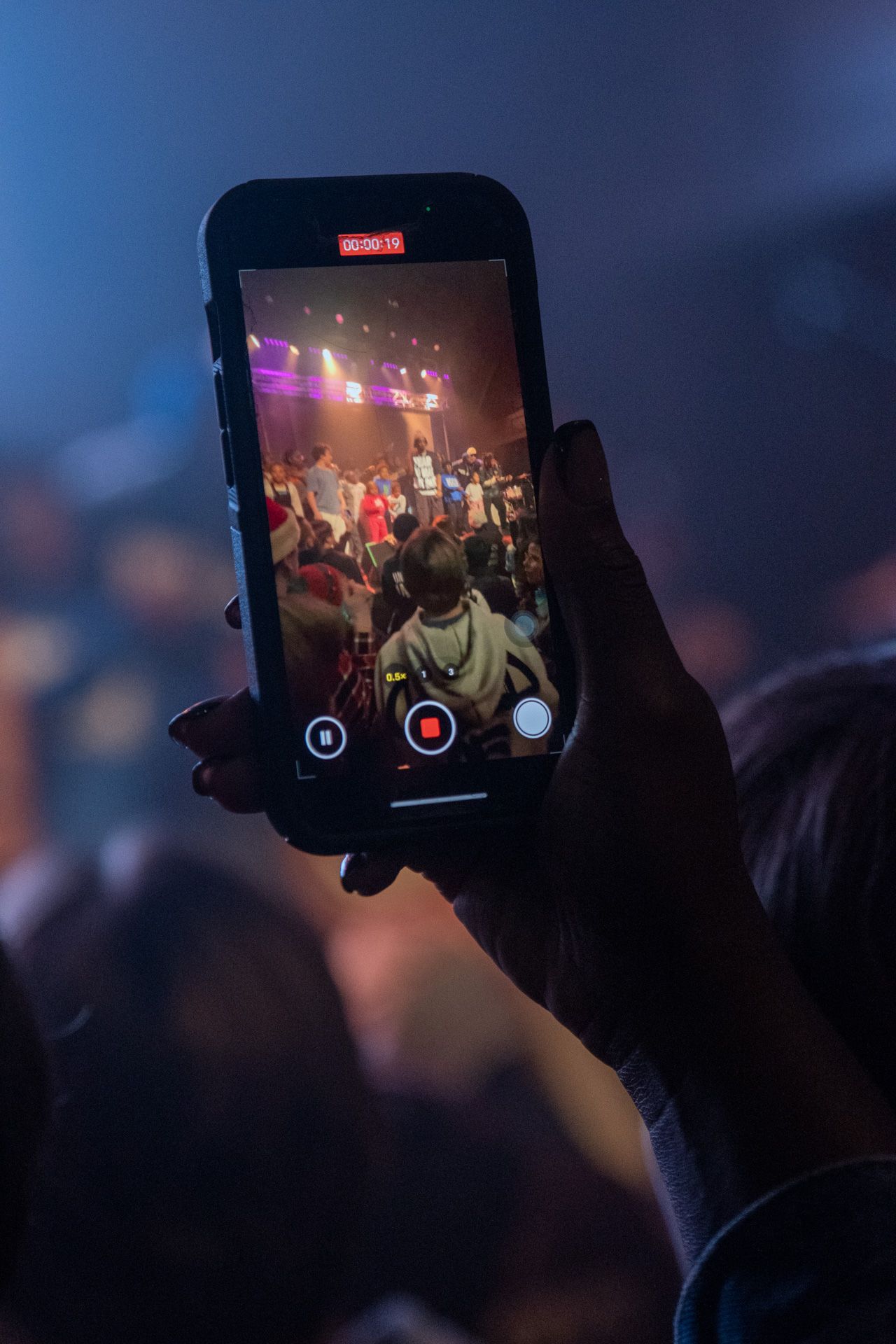 Silhouetted hands hold up mobile phones to record a concert performance in a dark venue with colorful stage lighting.