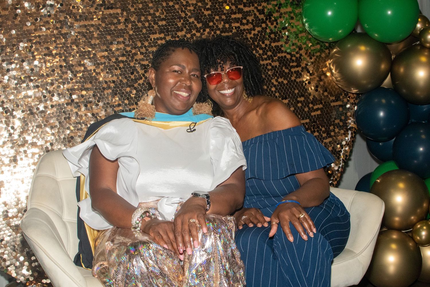 A joyful celebration with green and gold balloons in the background as two people embrace and smile warmly.