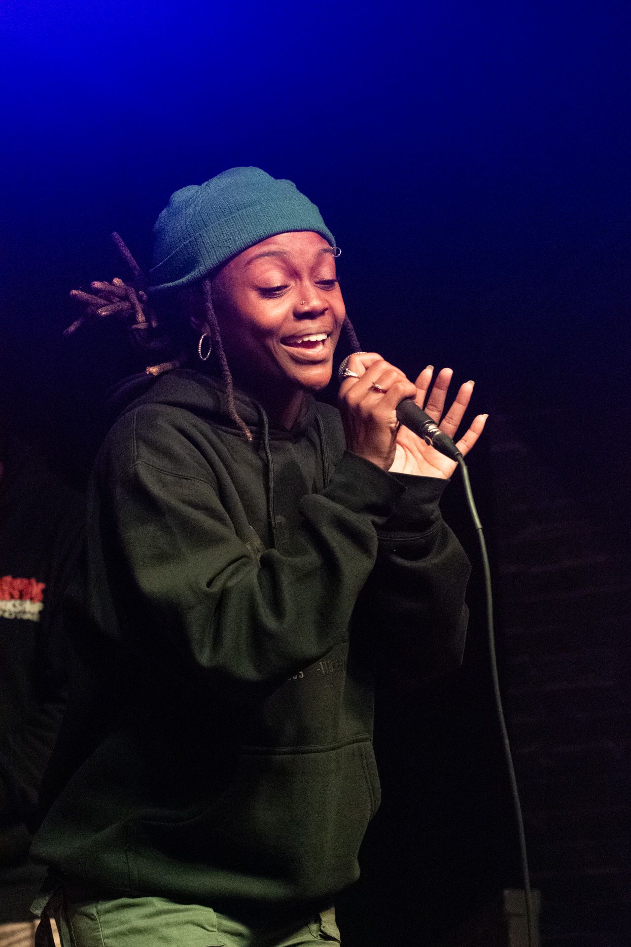 A performer in a green beanie and hoodie sings passionately into a microphone on a dark stage.