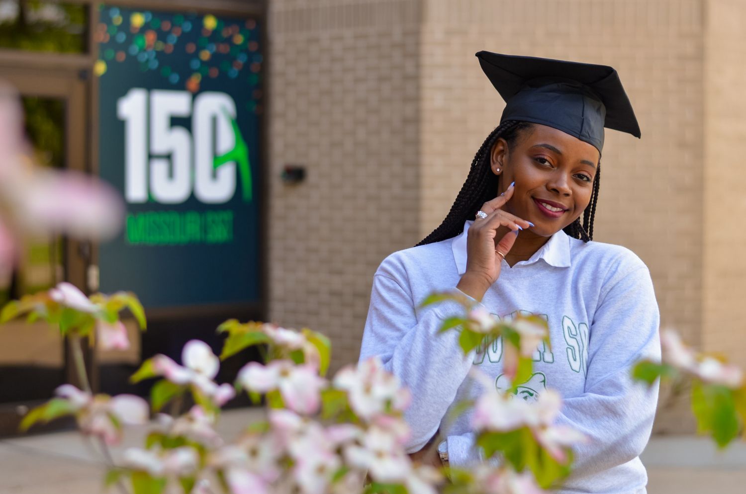 Black female college graduate smiles while in fornt of campus building with pink flowers in foreground.