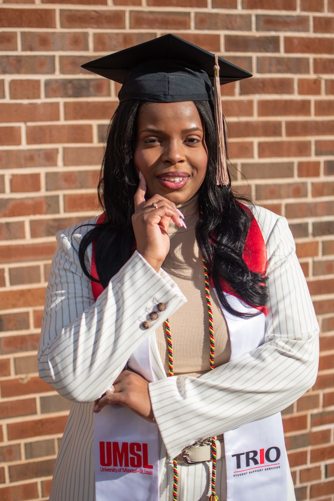Graduate in white sweater and graduation cap poses with diploma against brick wall.