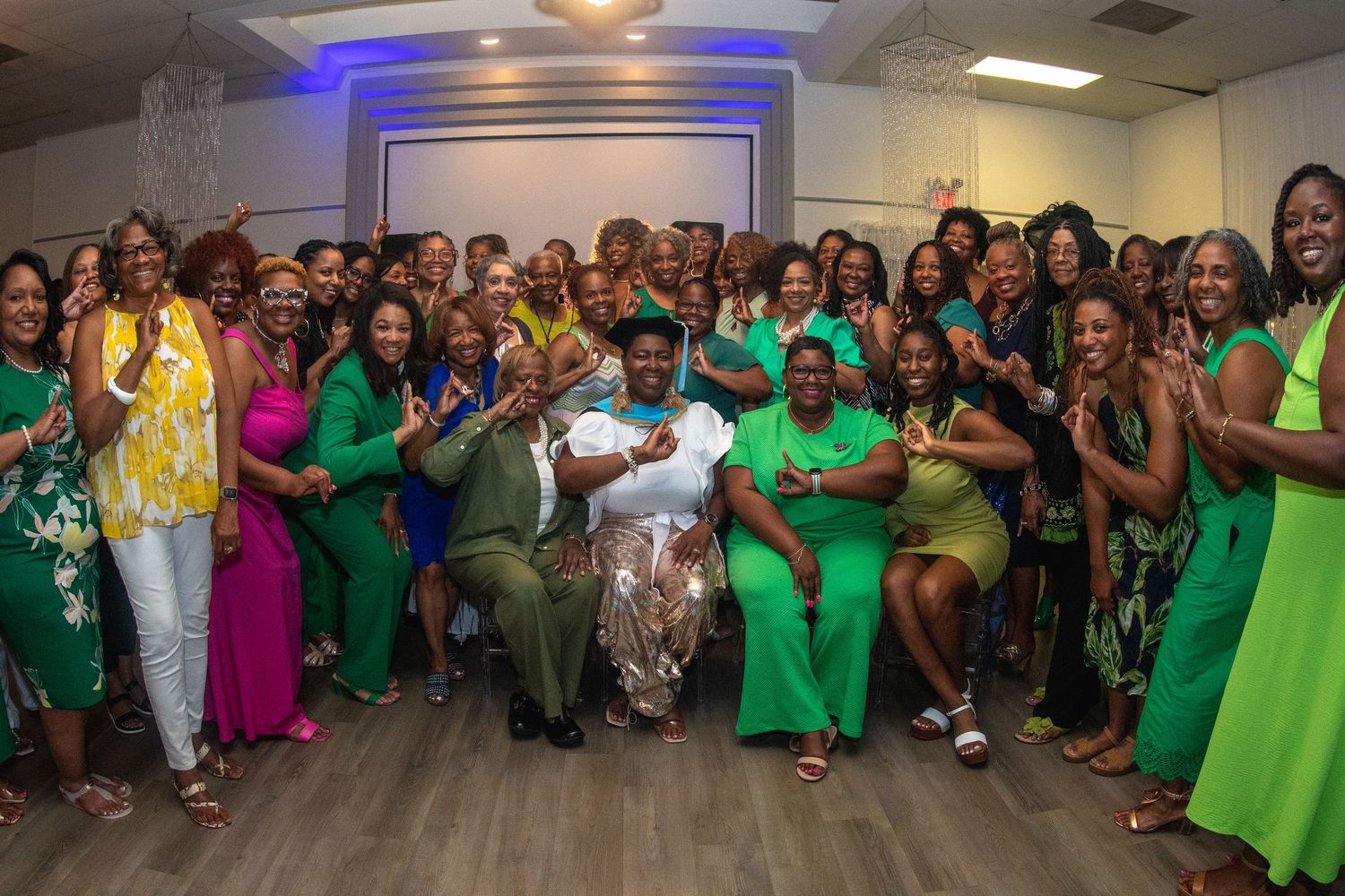 A large group of people wearing colorful green and pink outfits pose together for a group photo in an indoor setting.