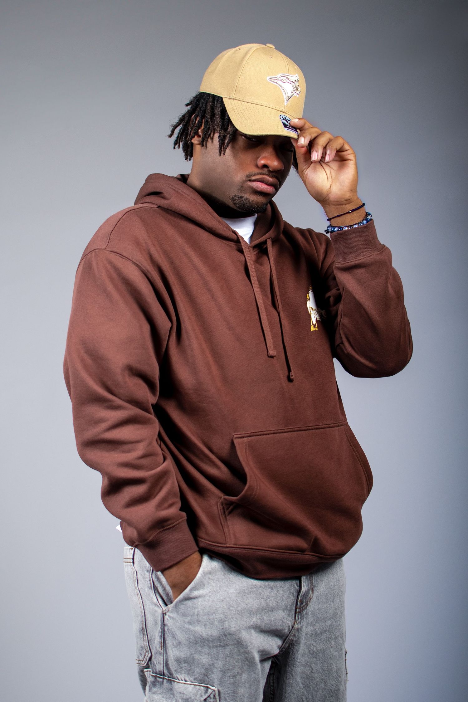 Person wearing brown hoodie and light jeans poses against gray background in fashion photography series.