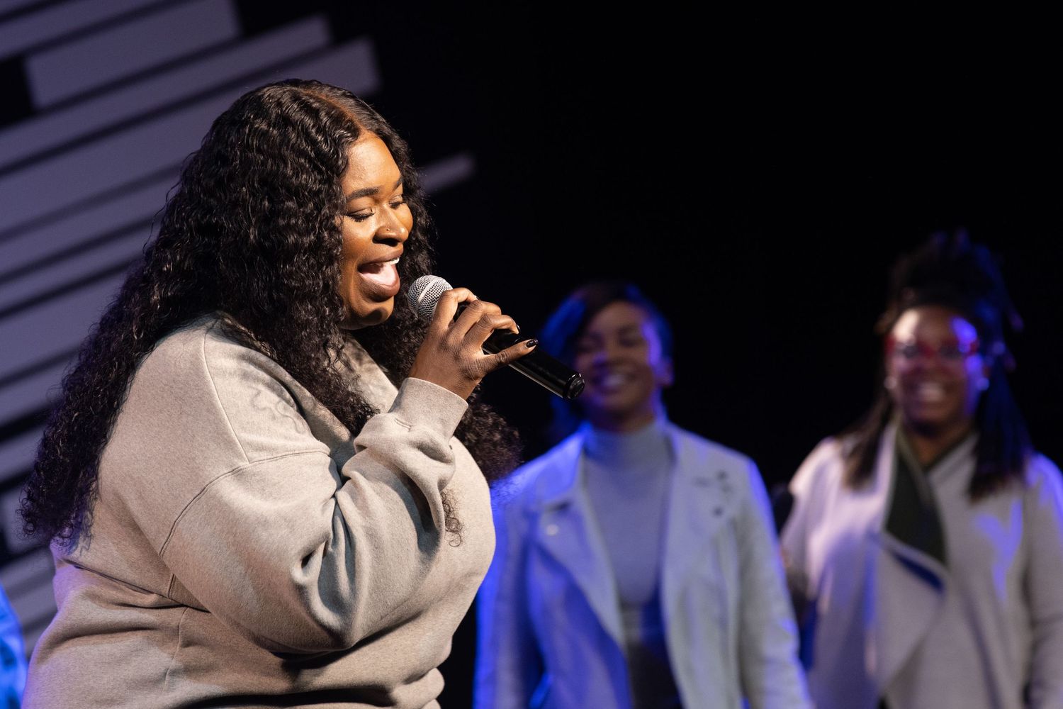 A performer in a white top sings passionately on stage with backup singers in blue outfits visible in the background.