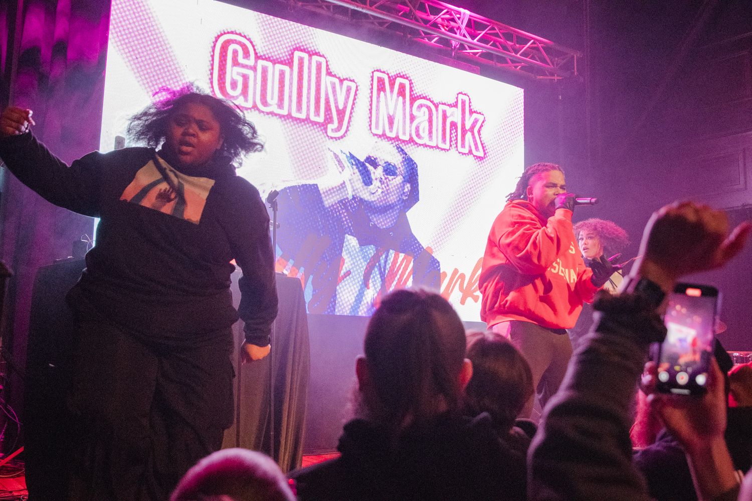 Concert performers on stage with vibrant pink and purple lighting creating an energetic atmosphere during a live show.