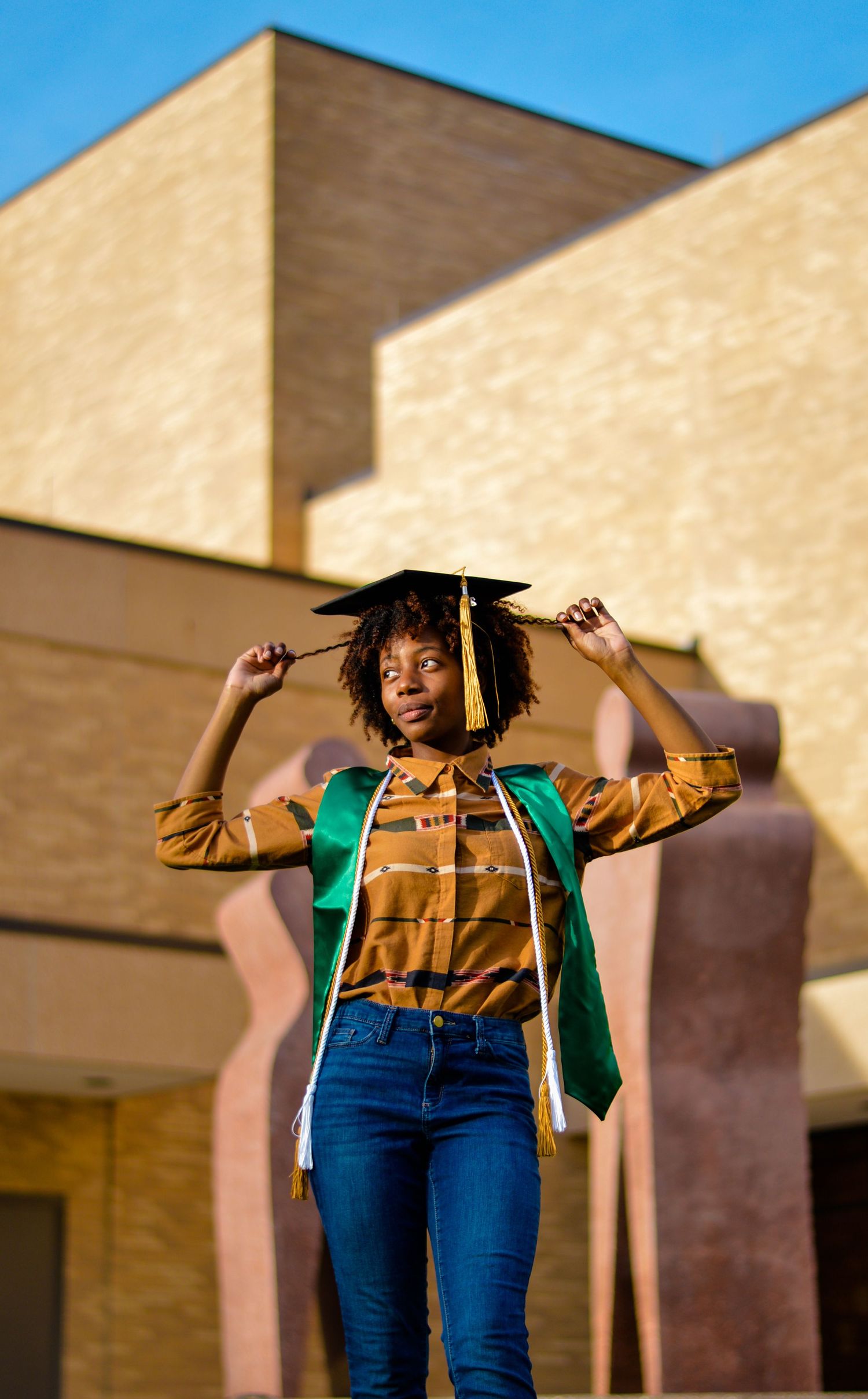 A graduate in blue jeans and gold top poses joyfully against a beige wall while wearing a graduation cap.