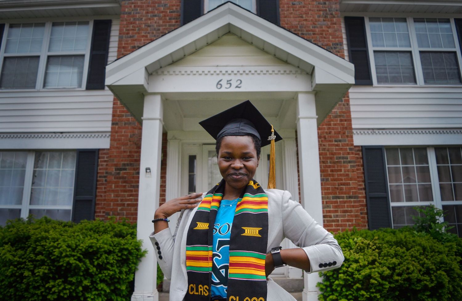 Graduate in white dress and kente stole poses in front of brick building entrance.