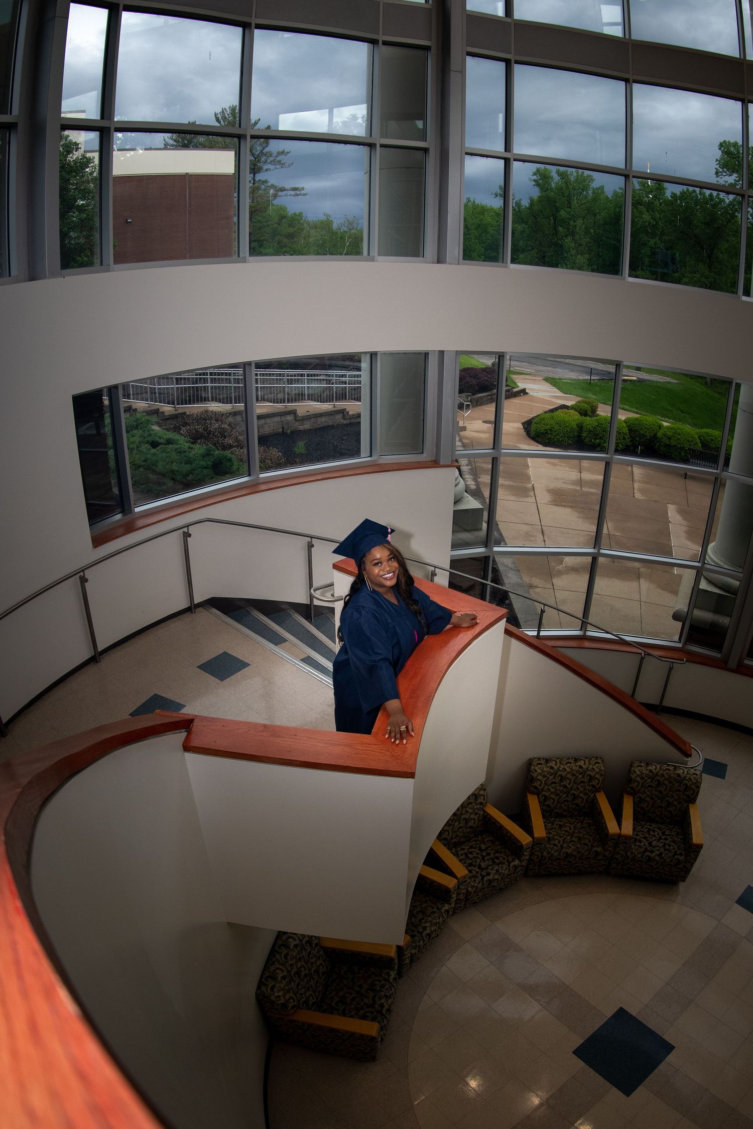 Graduate in blue cap and gown poses on curved modern staircase with large windows.