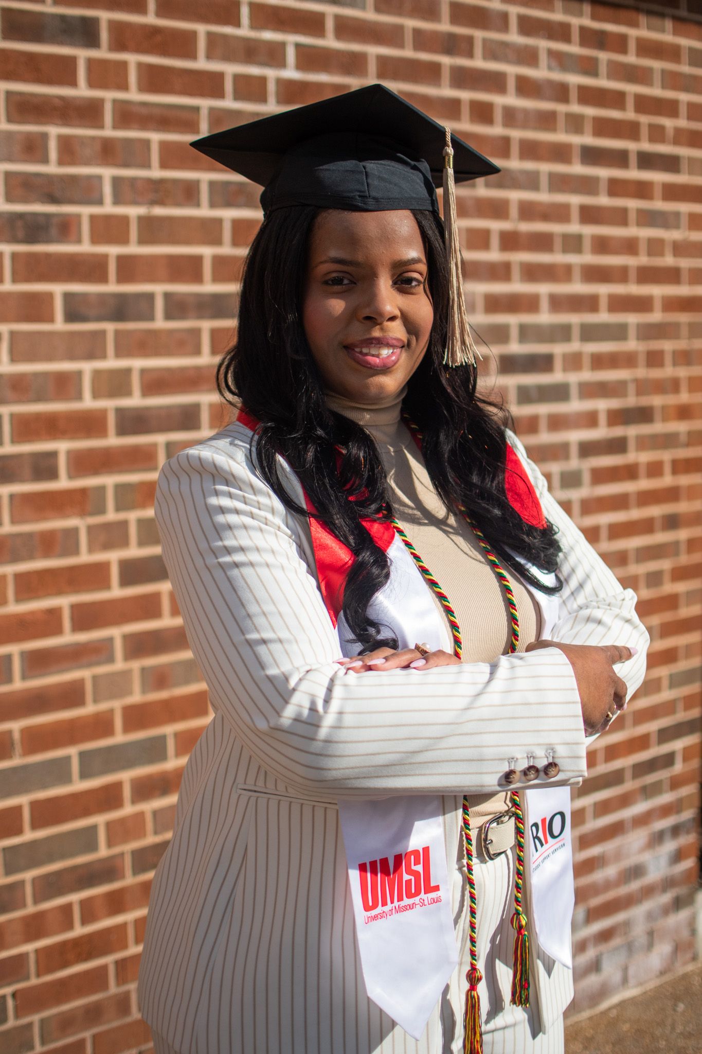 Graduate in cap and gown wearing white striped blazer and UMSL graduation stole poses against brick wall.