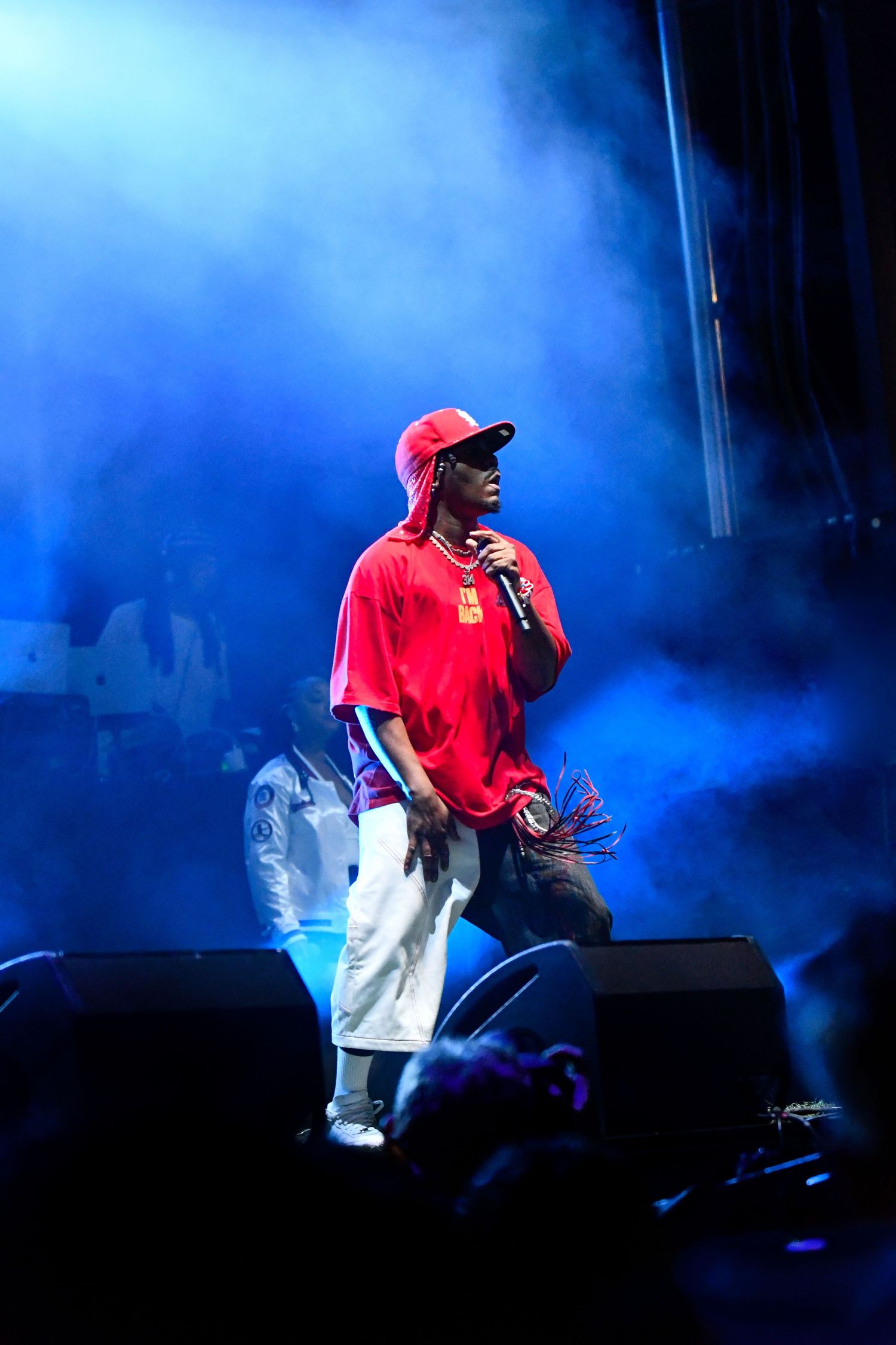 A performer in a red shirt and cap stands on stage illuminated by dramatic blue lighting and smoke effects.