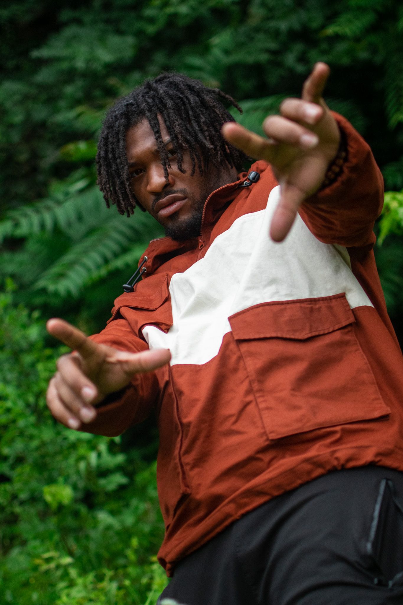 Person in a rust-colored and white jacket poses expressively against green foliage background.