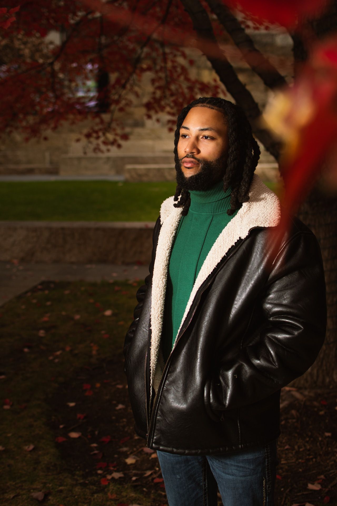 A person in a black leather jacket with sherpa lining and green turtleneck poses outdoors in autumn.