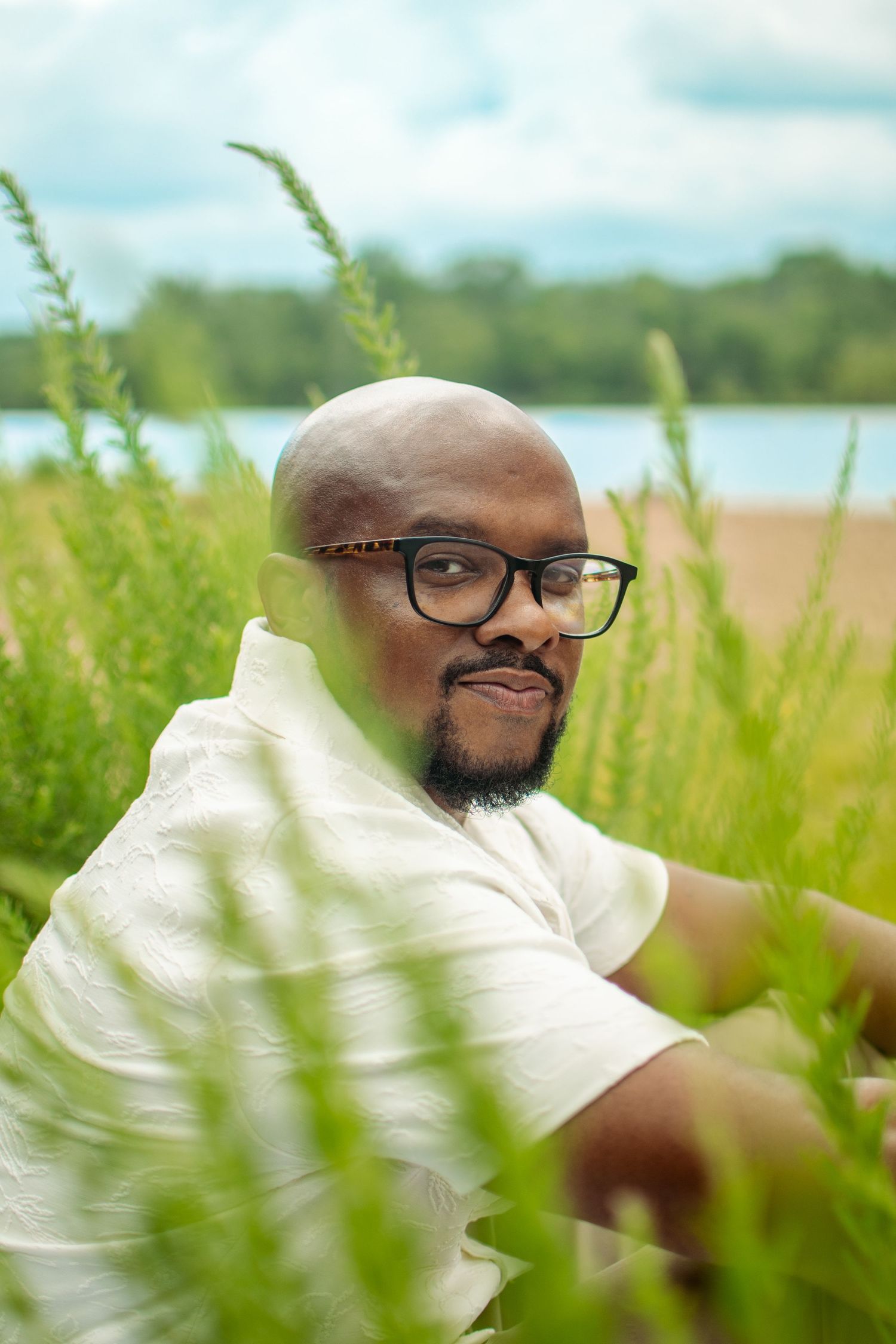 A person with glasses and a beard smiles while sitting in a lush green field on a sunny day.
