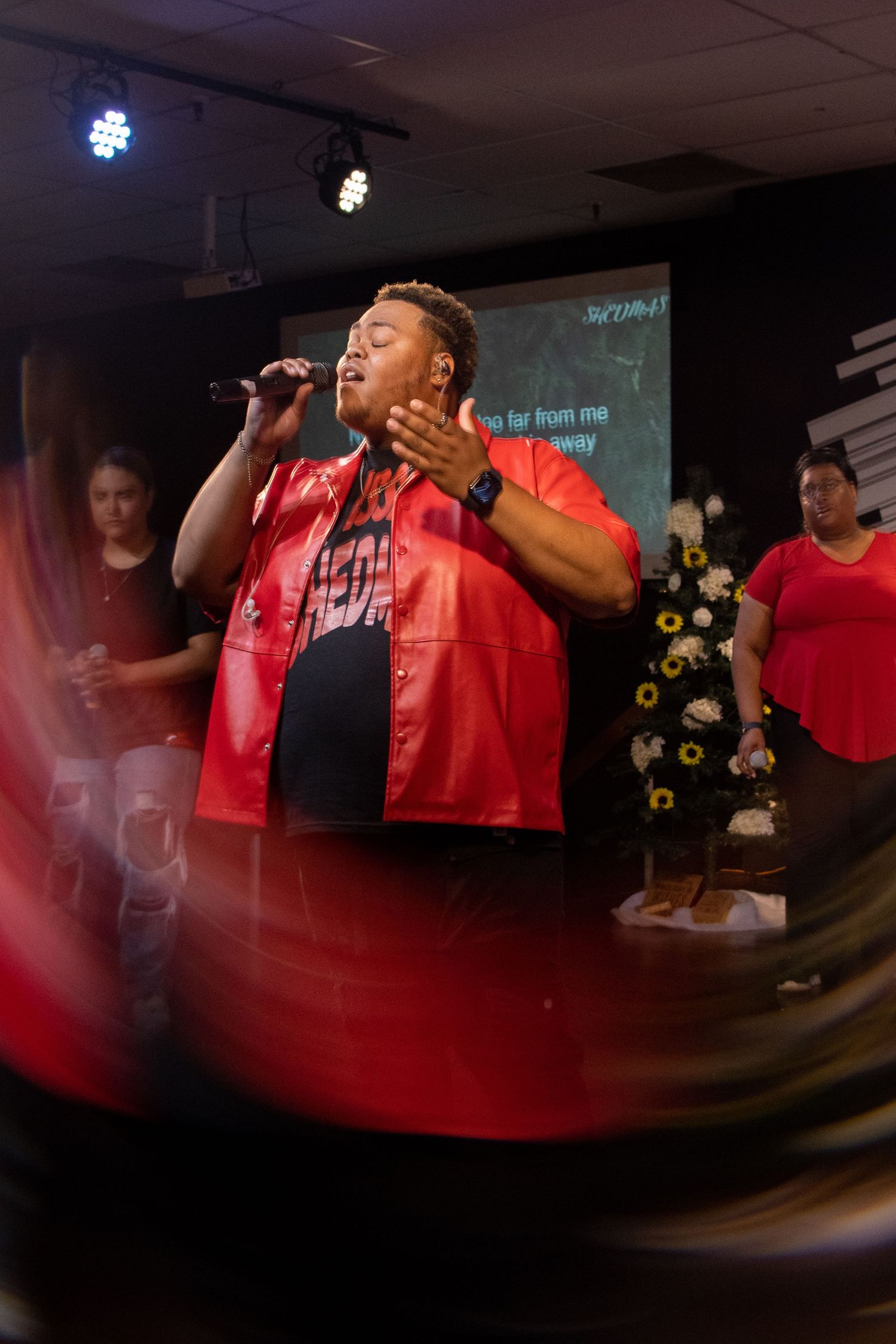 A performer in red clothing sings on stage in a dimly lit venue with dramatic lighting effects.