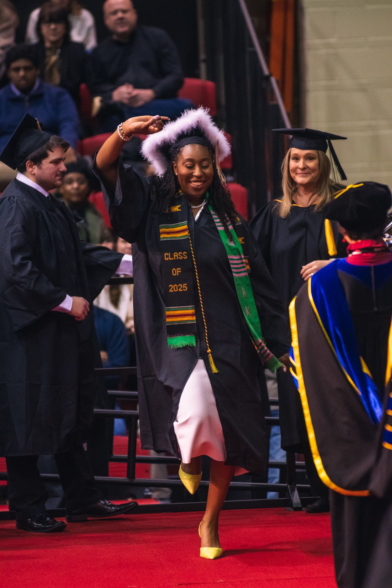 Students wearing graduation regalia and decorative stoles walk across a stage during graduation.