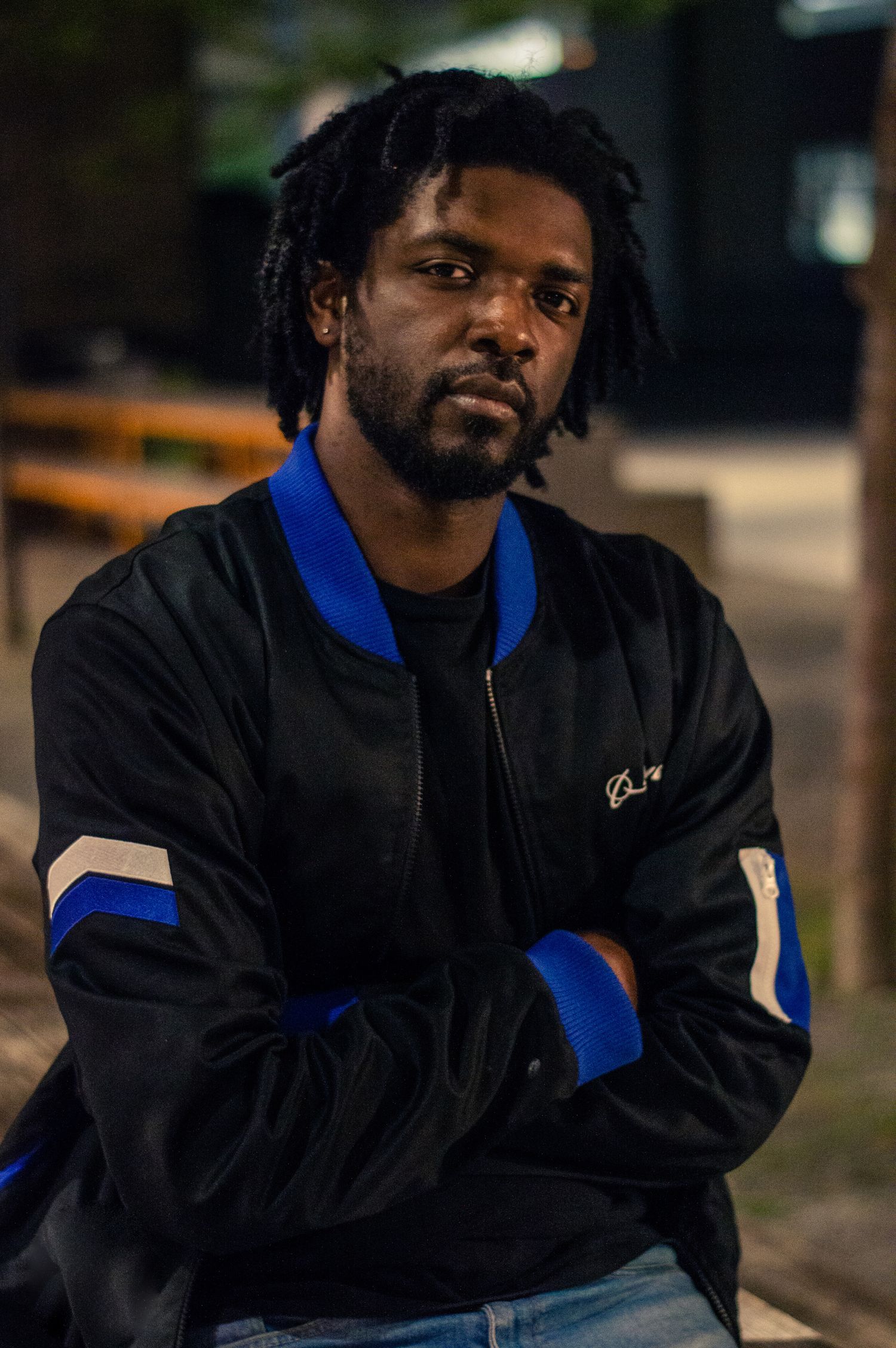 A black man wearing black jacket with blue accents sits in contemplative pose against blurred background.