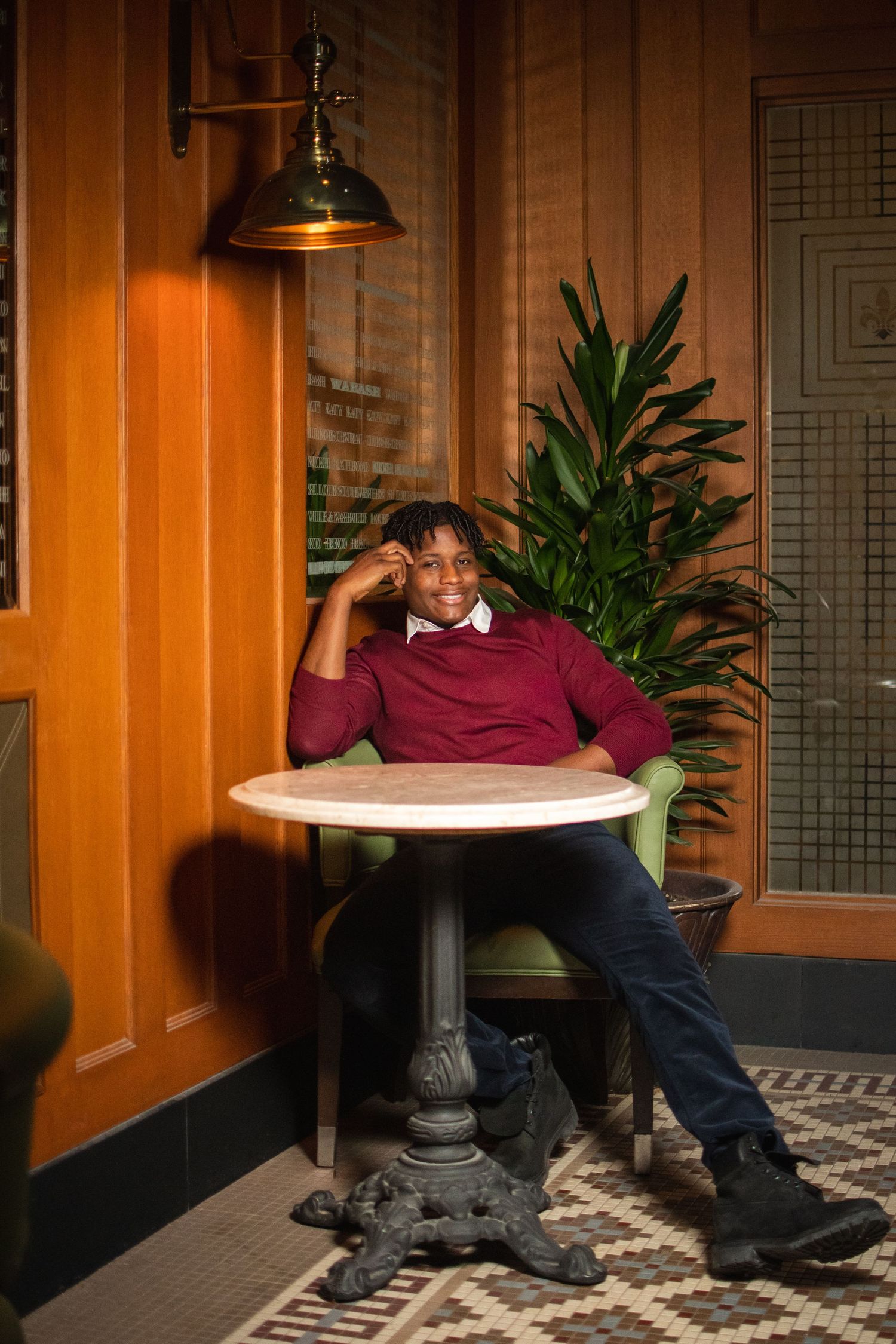 A person relaxes at a round bistro table with ornate base, wearing a burgundy sweater in a warmly lit restaurant setting.