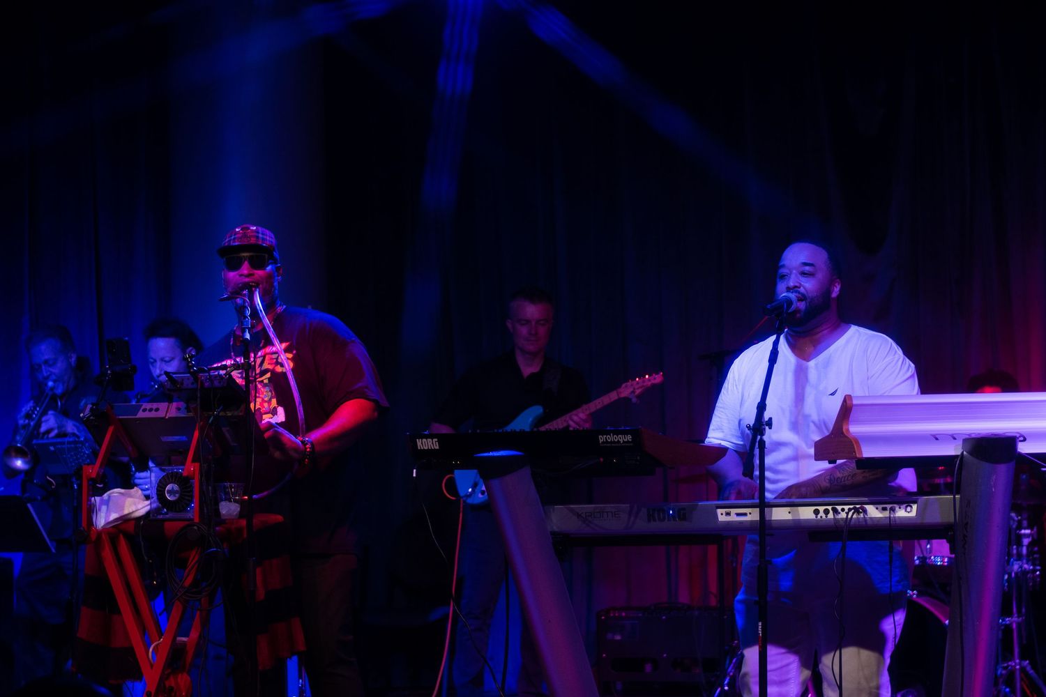 Musicians perform on a dark stage with dramatic red and blue lighting during a live concert performance.