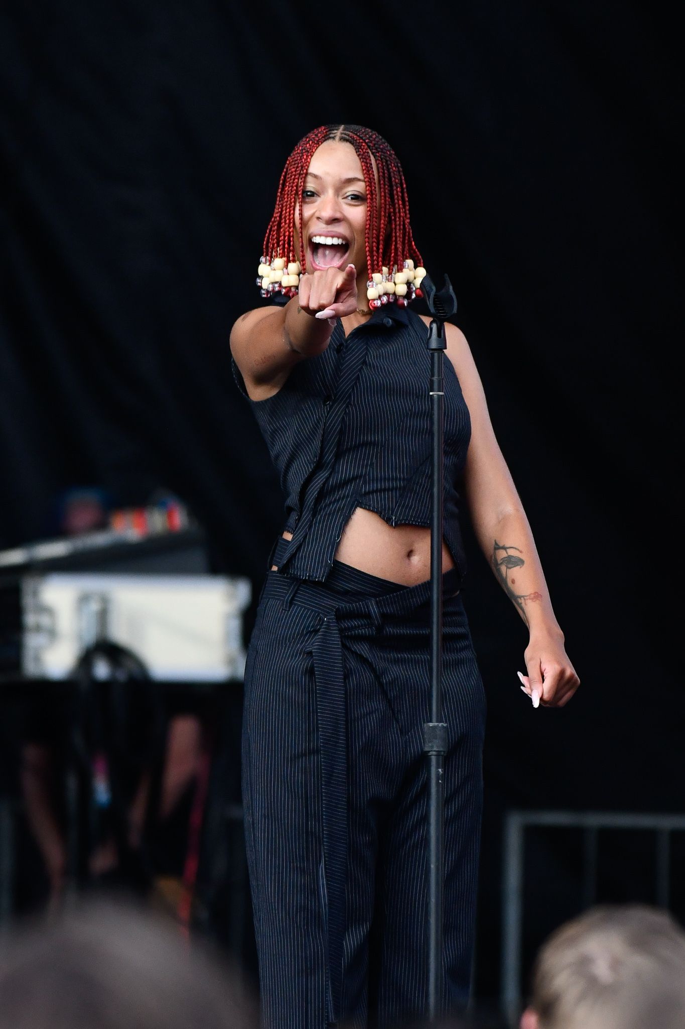 Performer in black crop top and pants singing energetically on stage with red braided hair and raised hands.