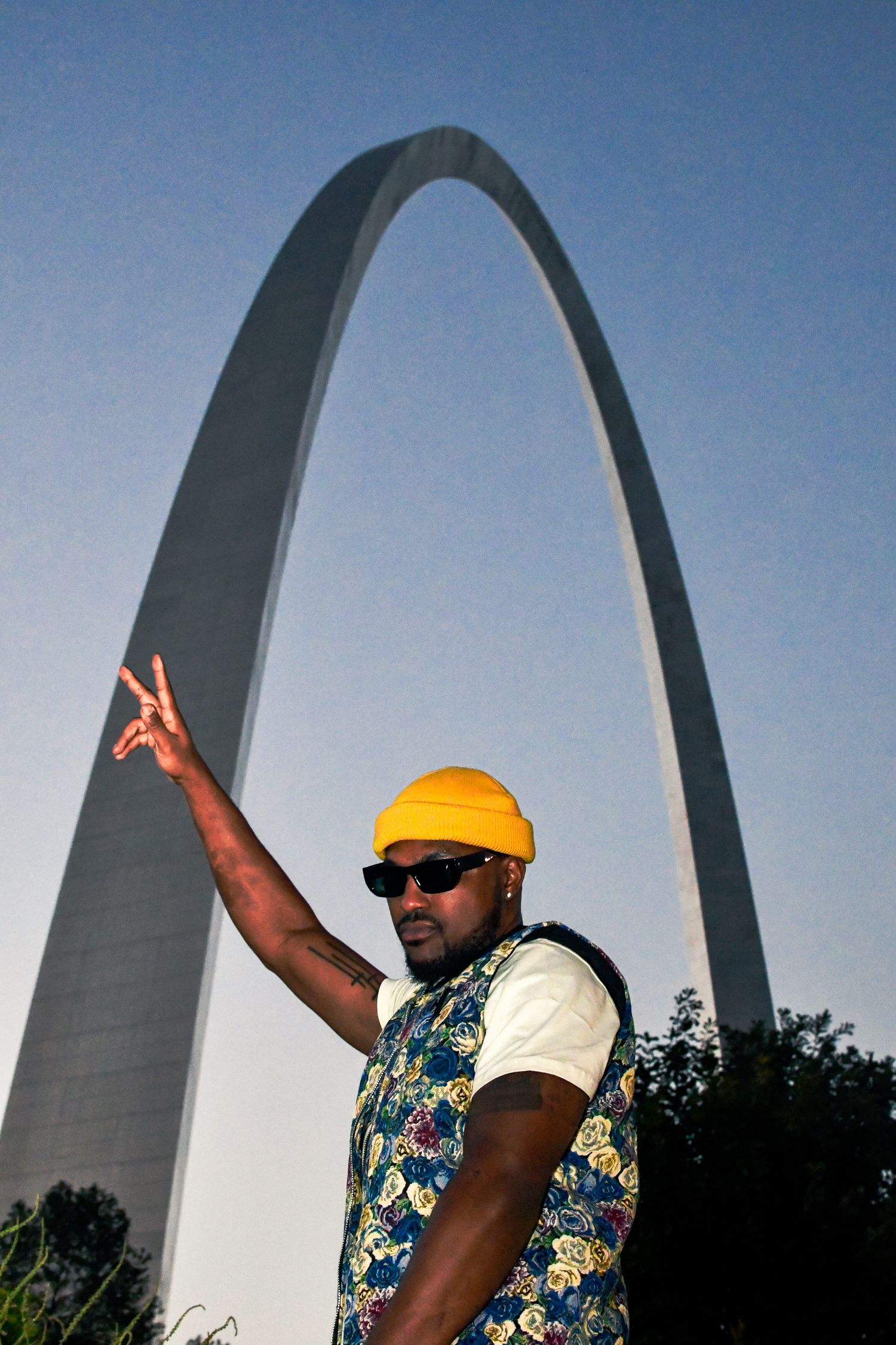 Multiple views of a person in a floral shirt and yellow hat posing in front of the Gateway Arch in St. Louis.
