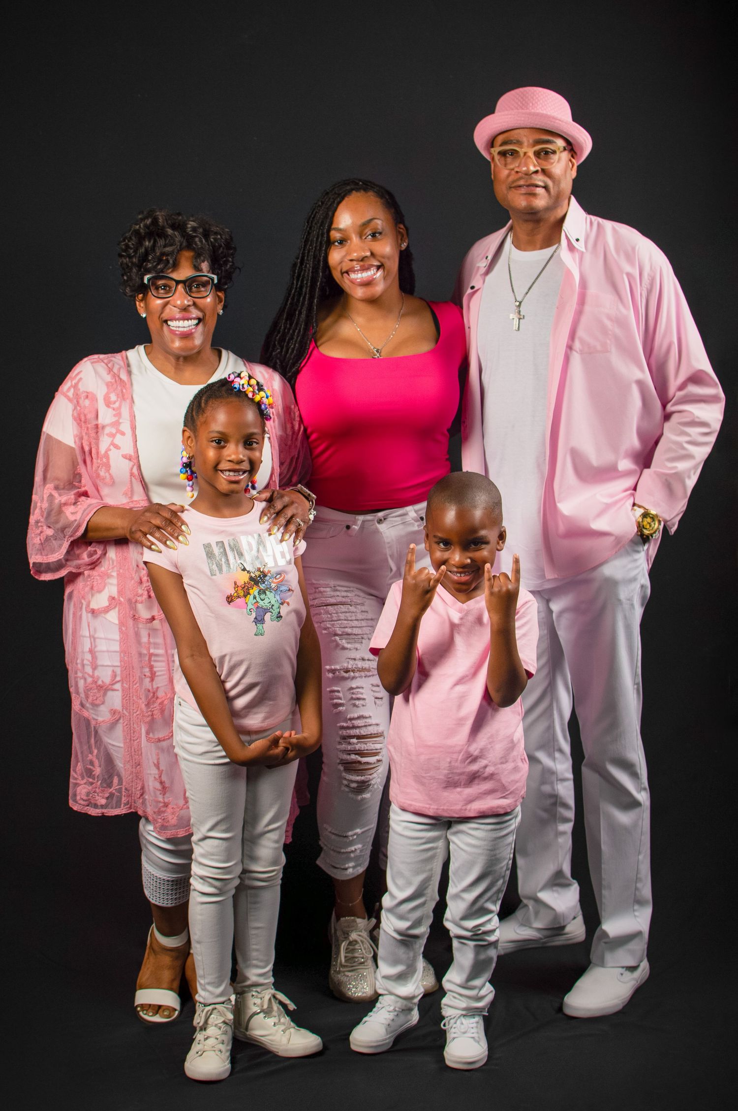 A family wearing coordinated pink and white outfits poses together against a black background in a studio setting.