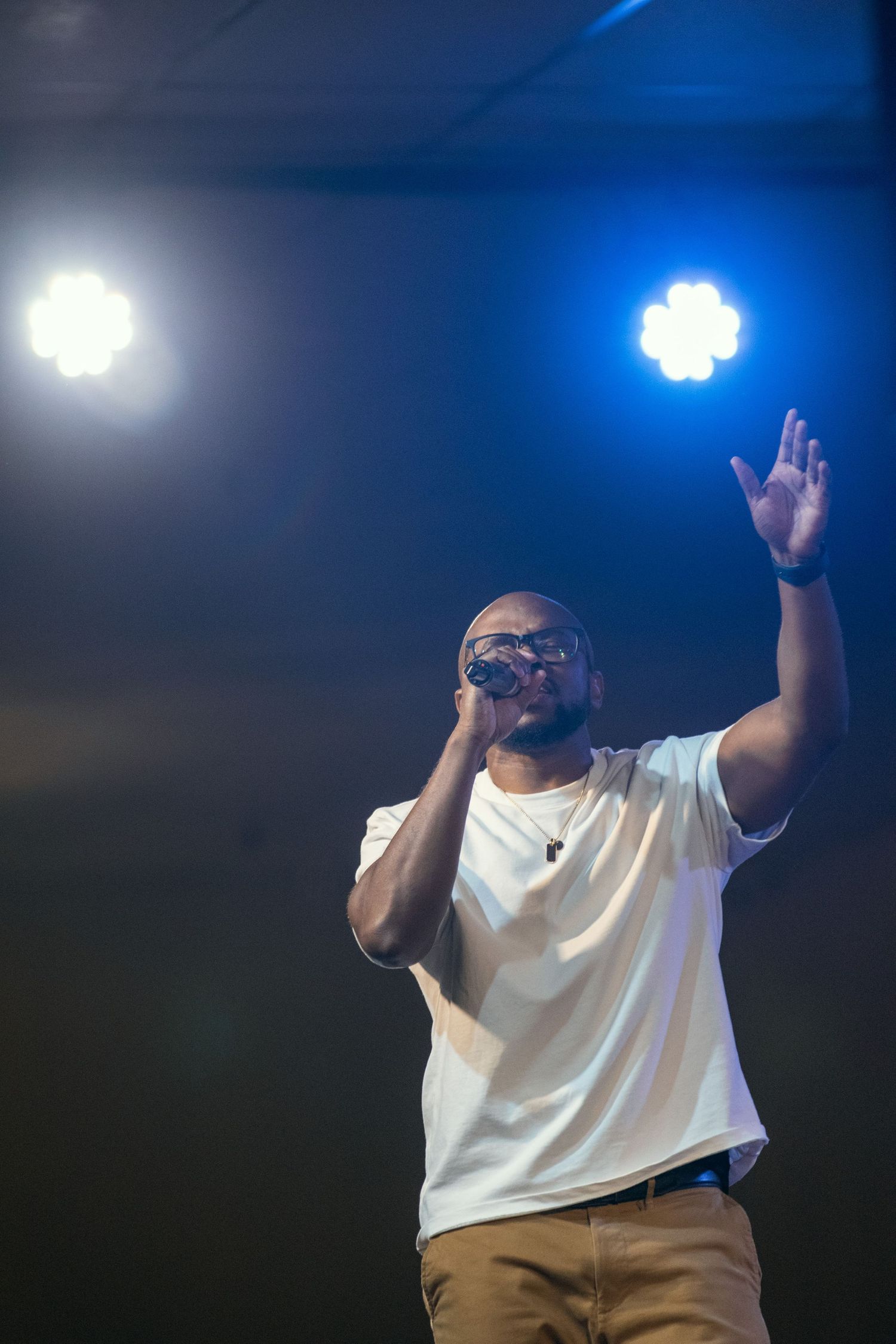 A performer in a white t-shirt and glasses raises their hands while on stage with blue lighting in the background.
