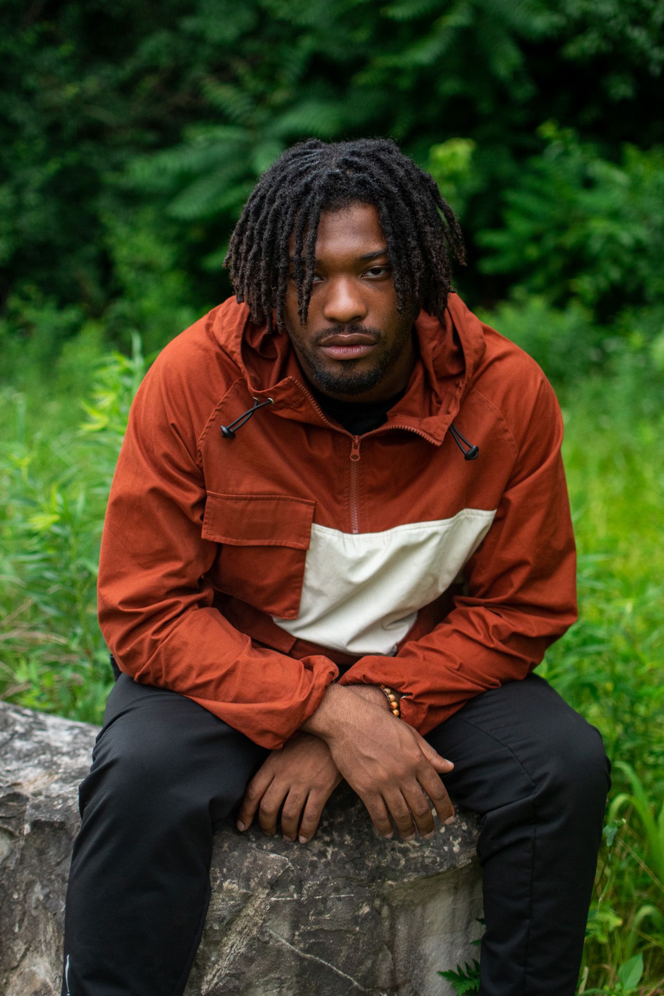 Person in rust-colored jacket sitting thoughtfully on stone wall surrounded by lush green foliage.